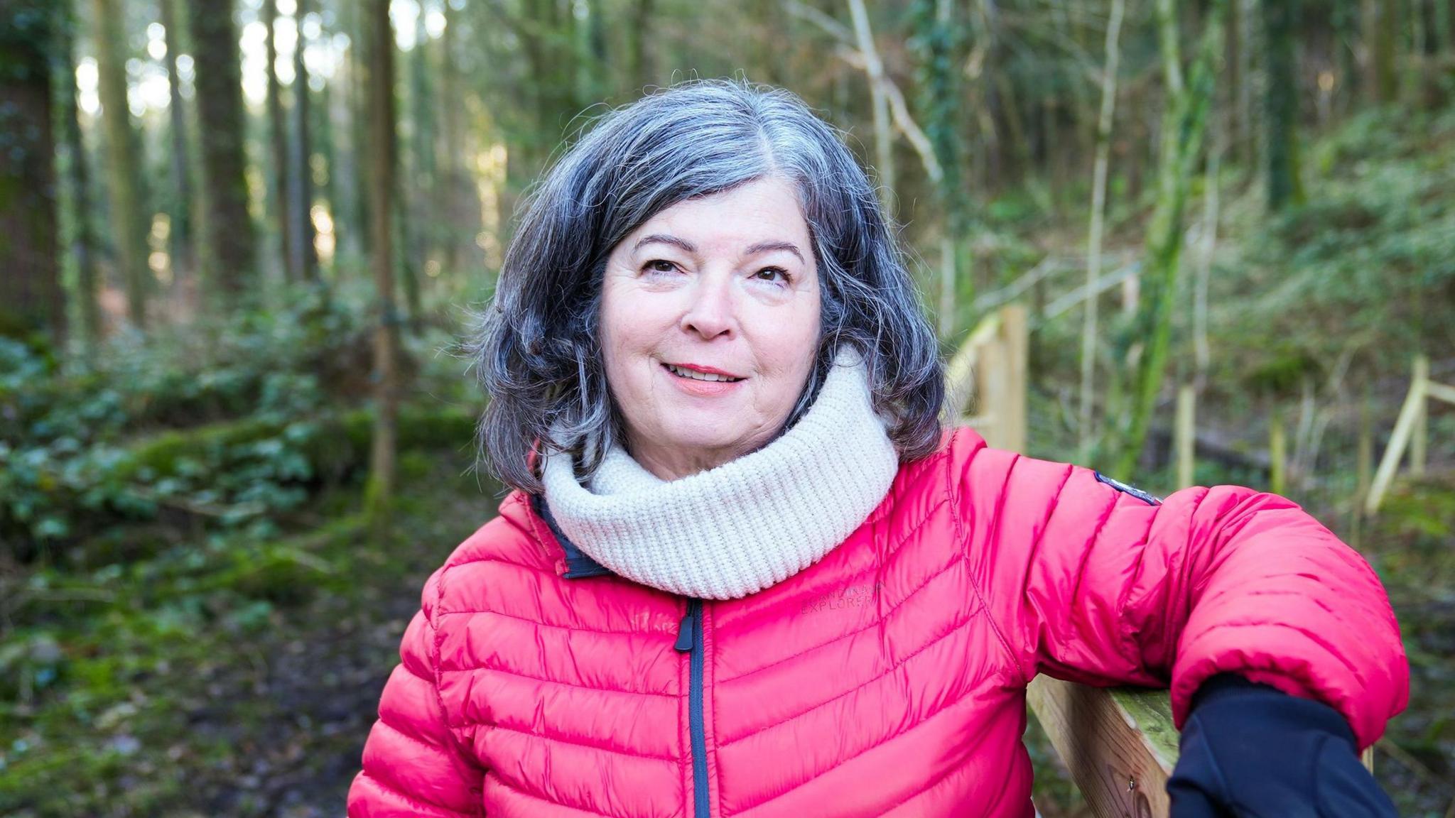 Ena Lloyd looking at the camera. She is black hair with wispy strands of grey. She is wearing a pink puffer coat and a white scarf. She is leaning on a wooden fence on the right of the picture. She is stood outside in woodland.