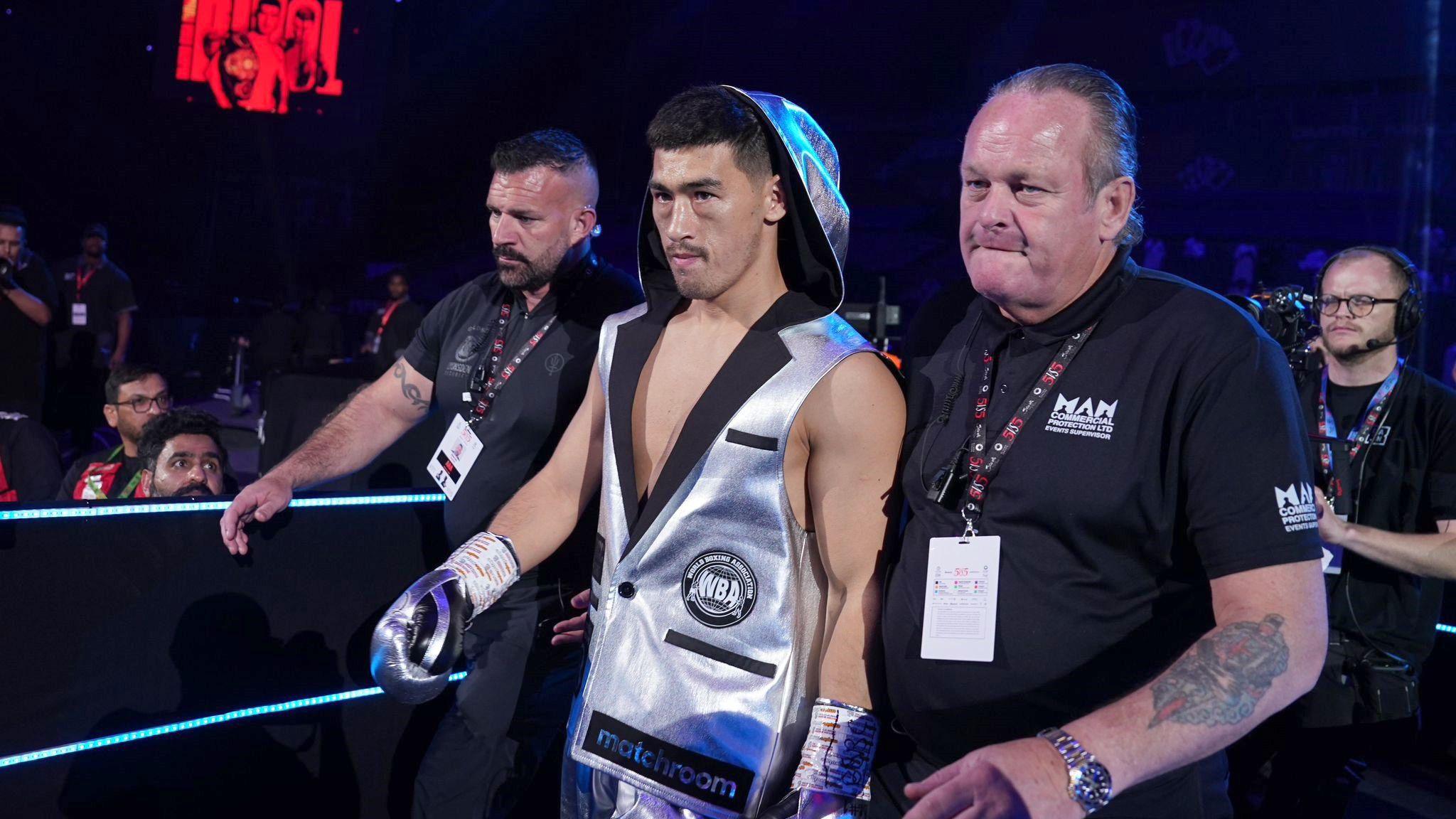 A boxer wearing a grey, hooded sleeveless jacket heading into the ring flanked by two security guards wearing back tops