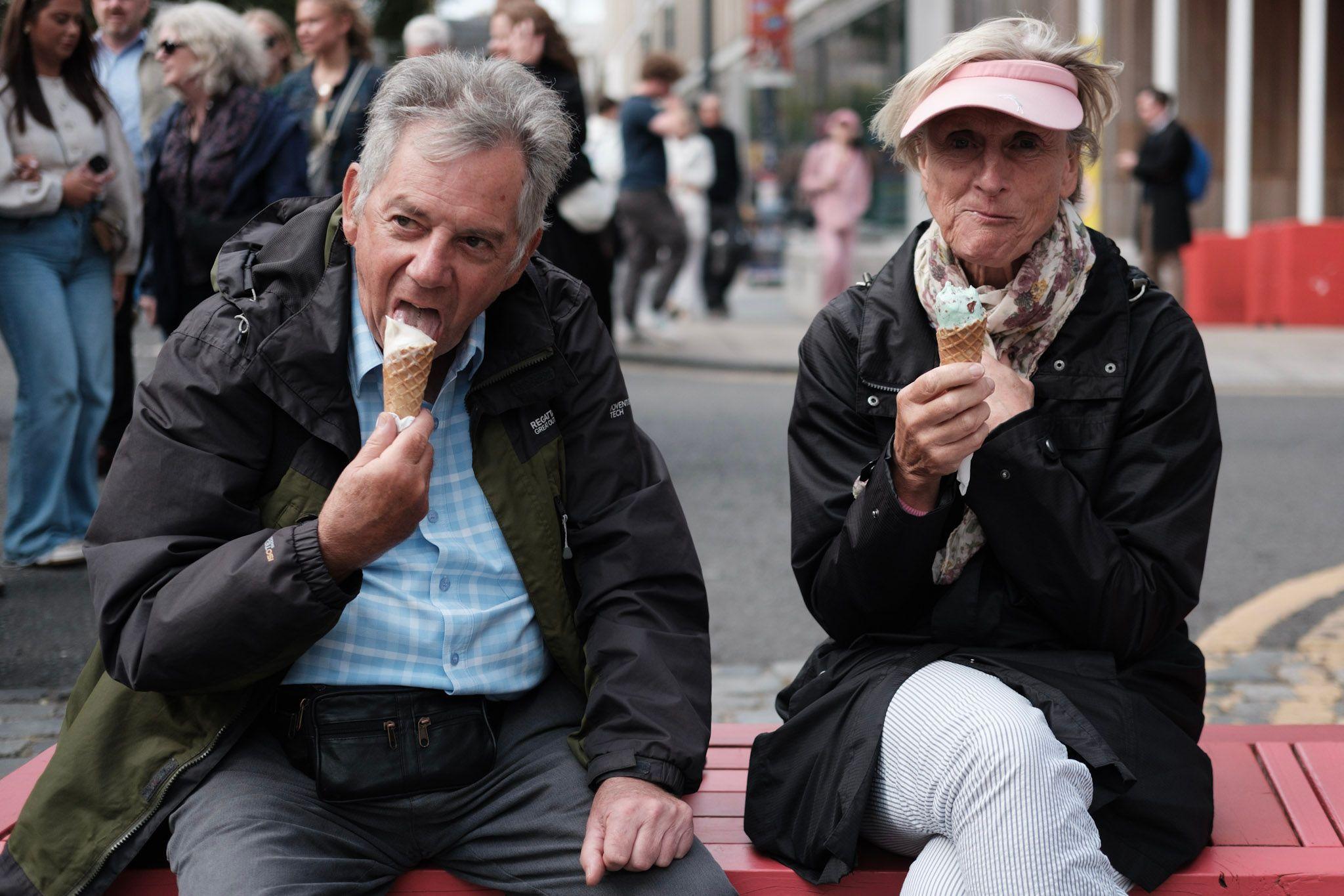 A middle-aged man and a woman sit on a street bench enjoying a couple of ice cream cones as the crowds pass by. The man wears a rain jacket and blue shirt as he takes a big lick of his cone, the woman, wearing a pink visor cap holds her mint choc chip cone.