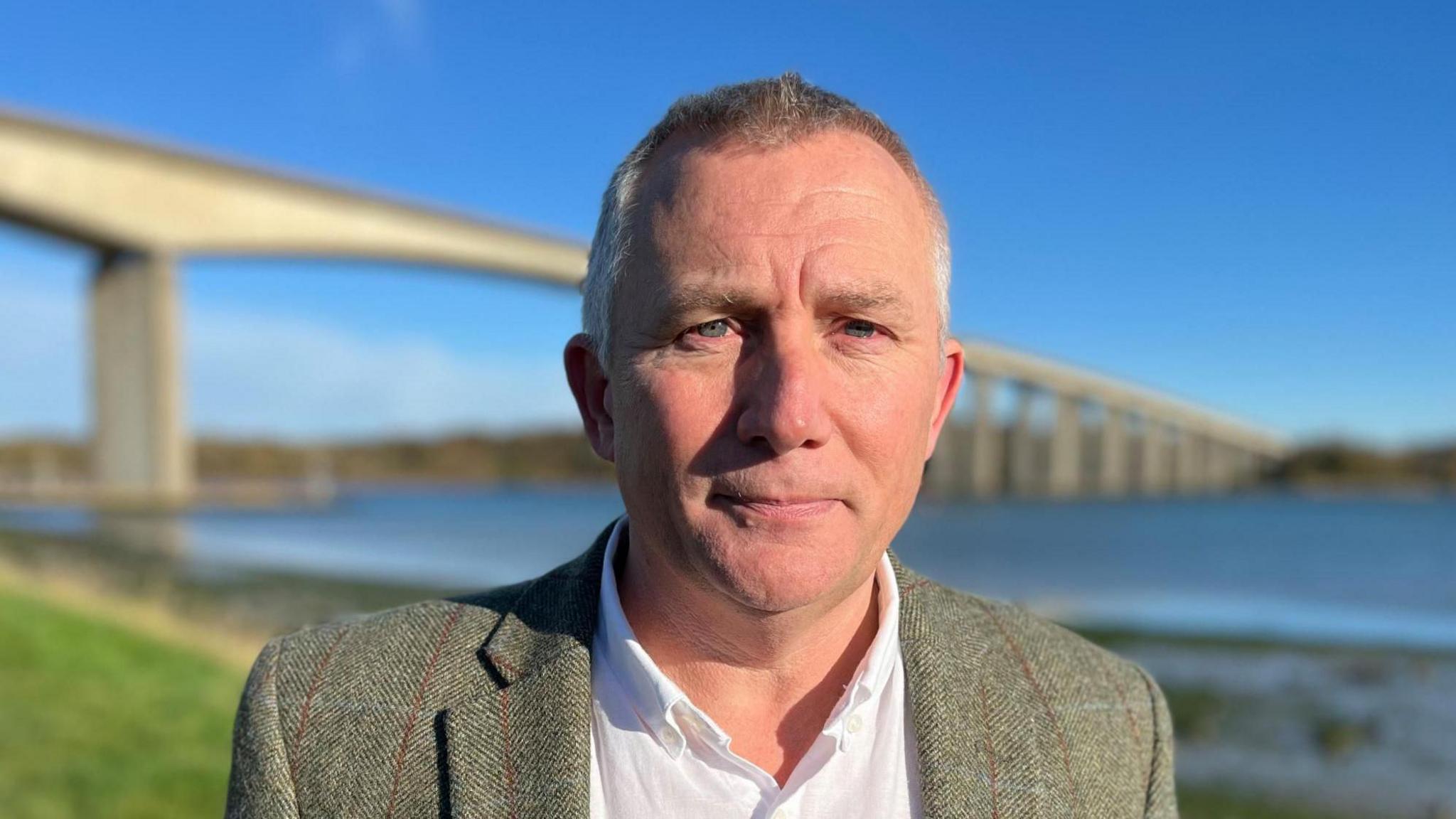 Mark Ling wearing a jacket and a white shirt standing in front of the Orwell Bridge and the River Orwell 