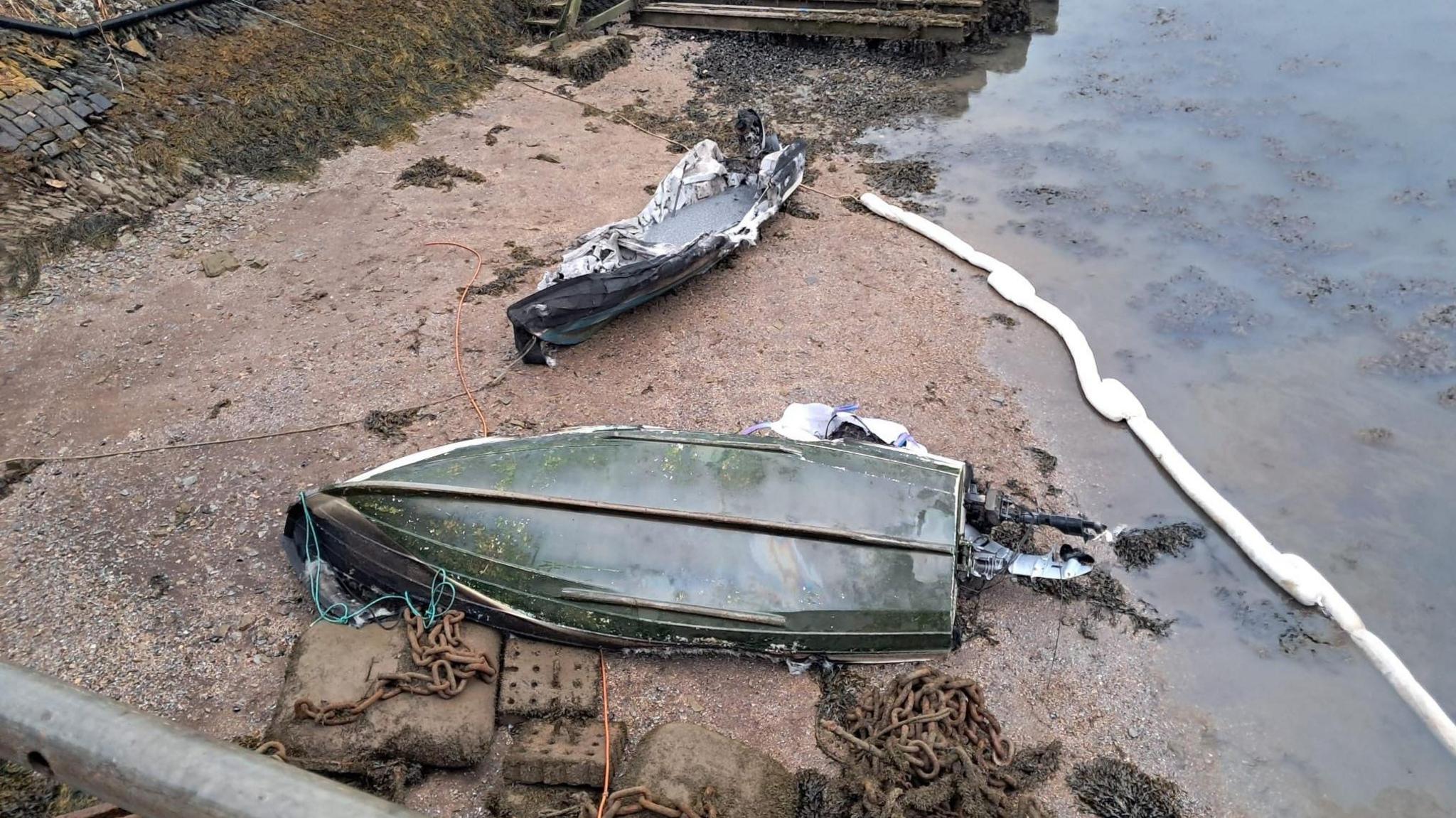 Two burned boats on sand. The boat in the bottom of picture is upside down. The boat in the tip of the picture is the right way up but appears to be melted in some parts. There is clear water with seaweed in it in the top right corner. 