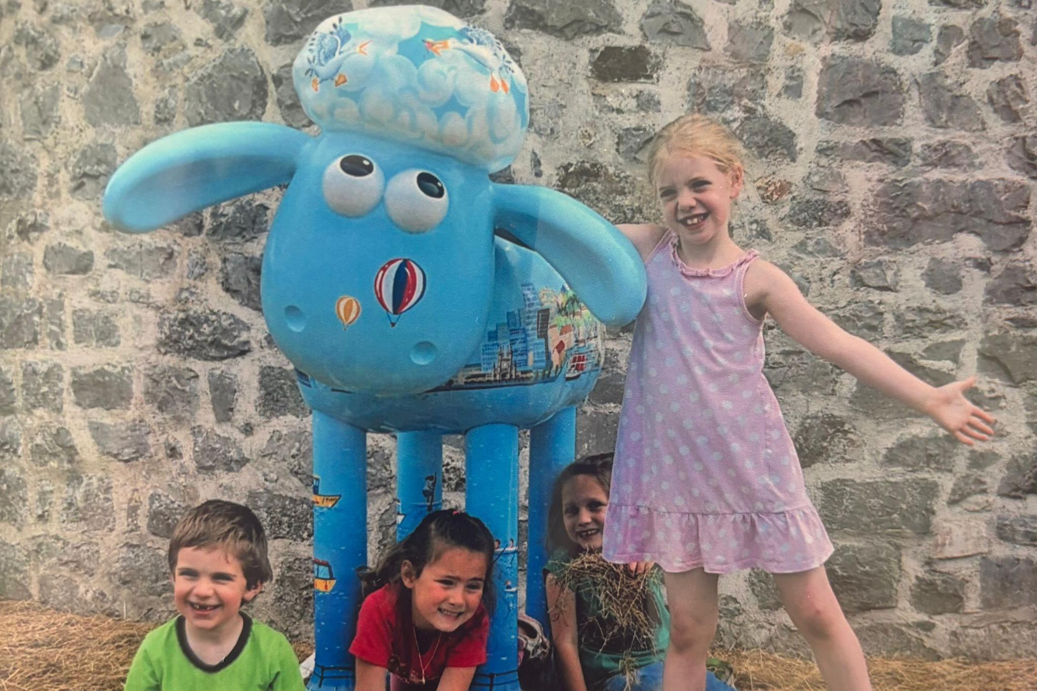 A younger Naomi, aged five, wears a pink dress and stands beside a pale blue Shaun the Sheep sculpture decorated with colourful hot air balloons on its face. Three other children — her siblings and a friend — are sitting under the sculpture, smiling for the camera.