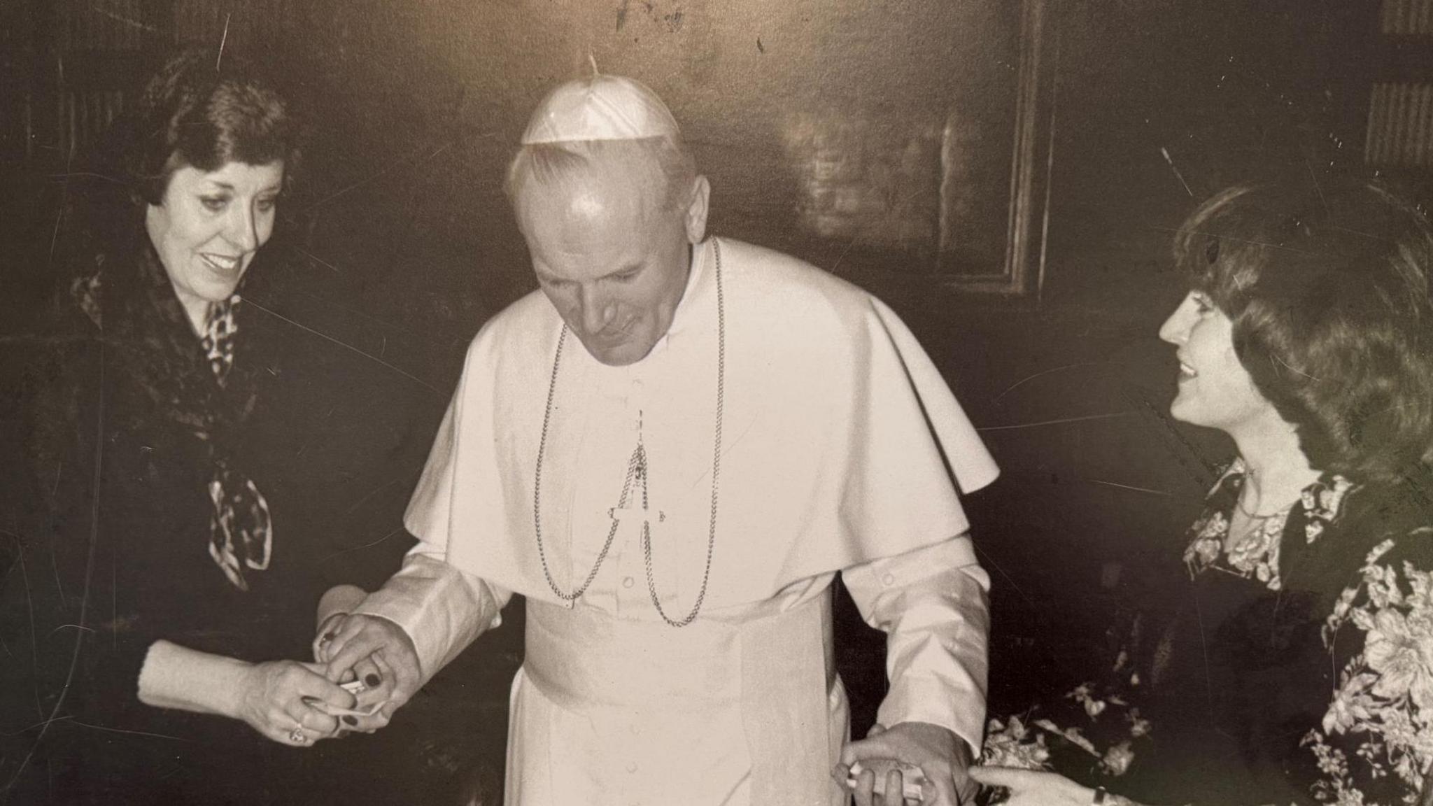 Two women with 1980s style hair hold either hand of Pope John Paul II, dressed in his white papal gown and skullcap. 