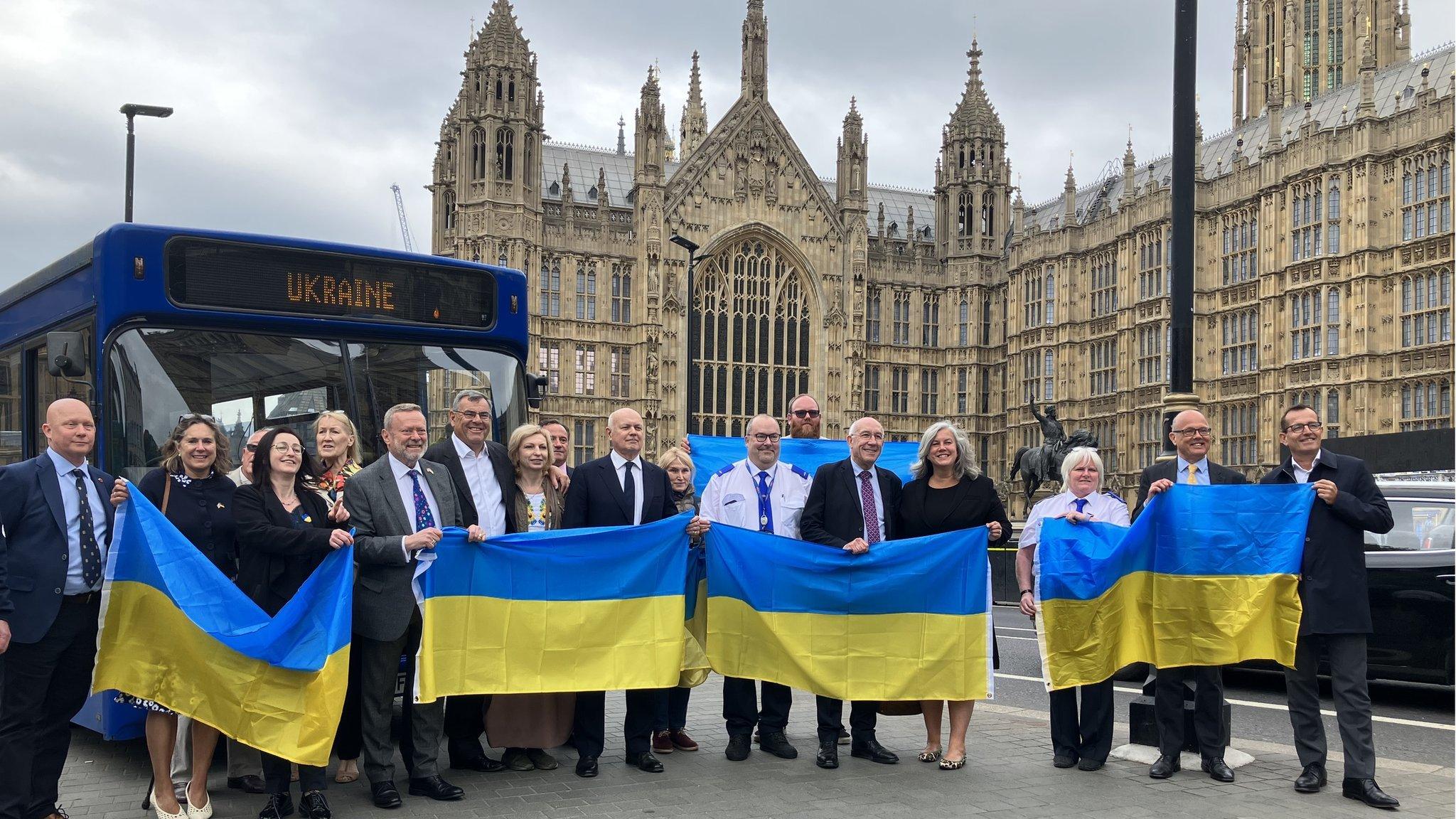 A group of people holding Ukrainian flags stand in front of a single decker bus that's been converted into a field hospital, outside the House of Lords in Westminster