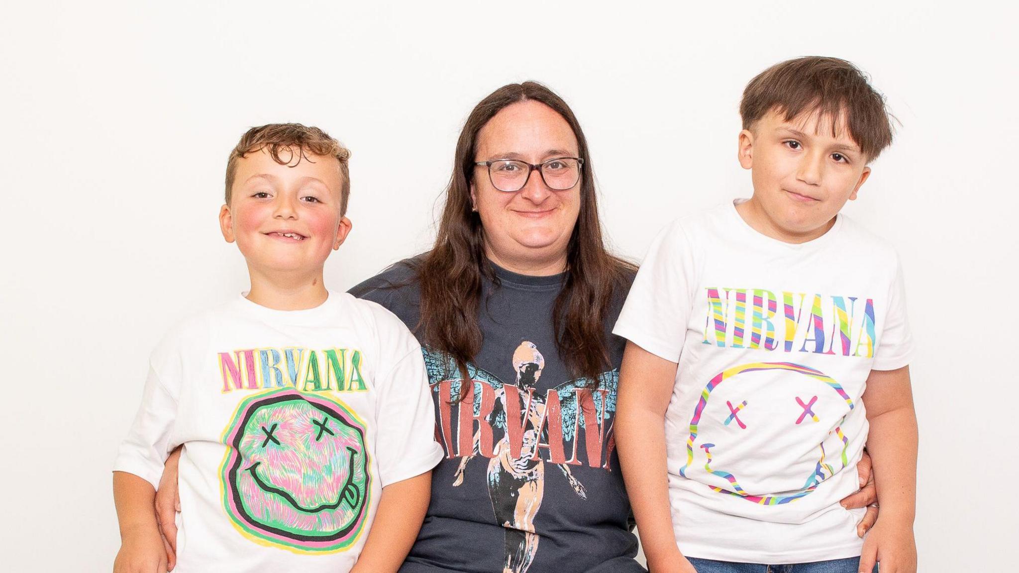 Two young boys with brown hair stand either side of a their mother. She has long brown hair, glasses and is wearing a grey Nirvana T-shirt with pink writing and a person with angel wings on it. Both boys are wearing white Nirvana T-shirts with a round smiley face with crosses for eyes. They are all looking at the camera and smiling. 