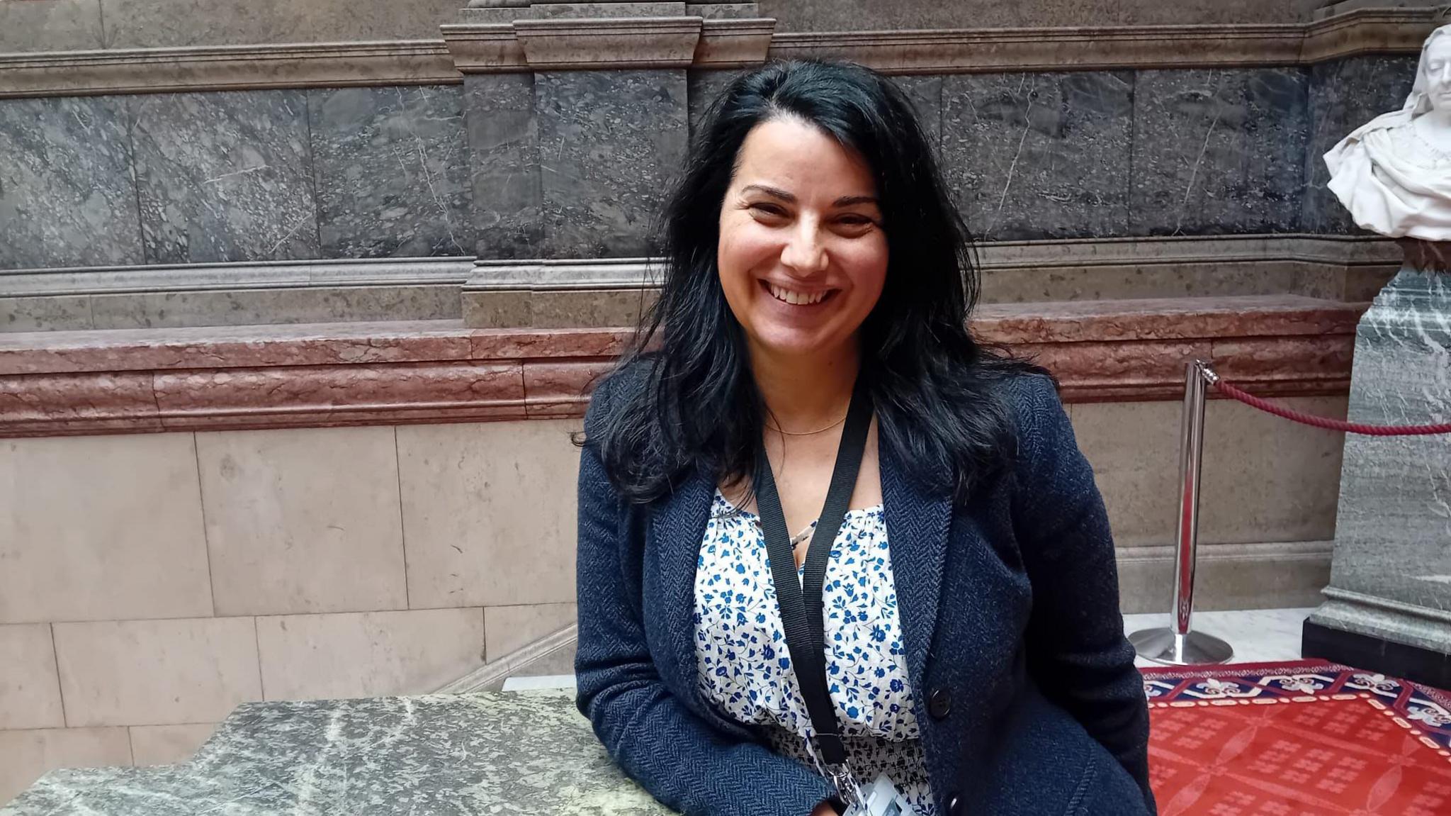 A woman with long black hair is wearing a navy jacket and a white dress with small blue flowers. She is stood on the marble staircase at Sheffield town hall and is smiling