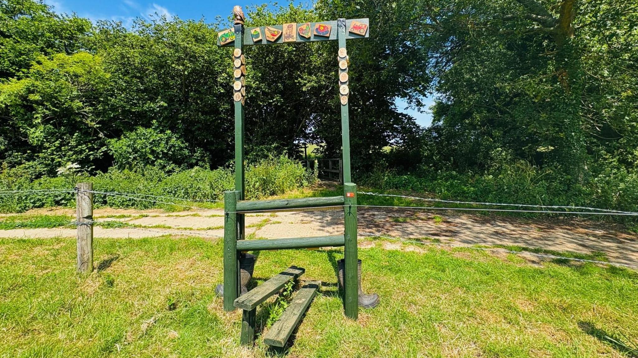 A stile over a wire fence, made of green posts with an archway decorated with painted wood blocks