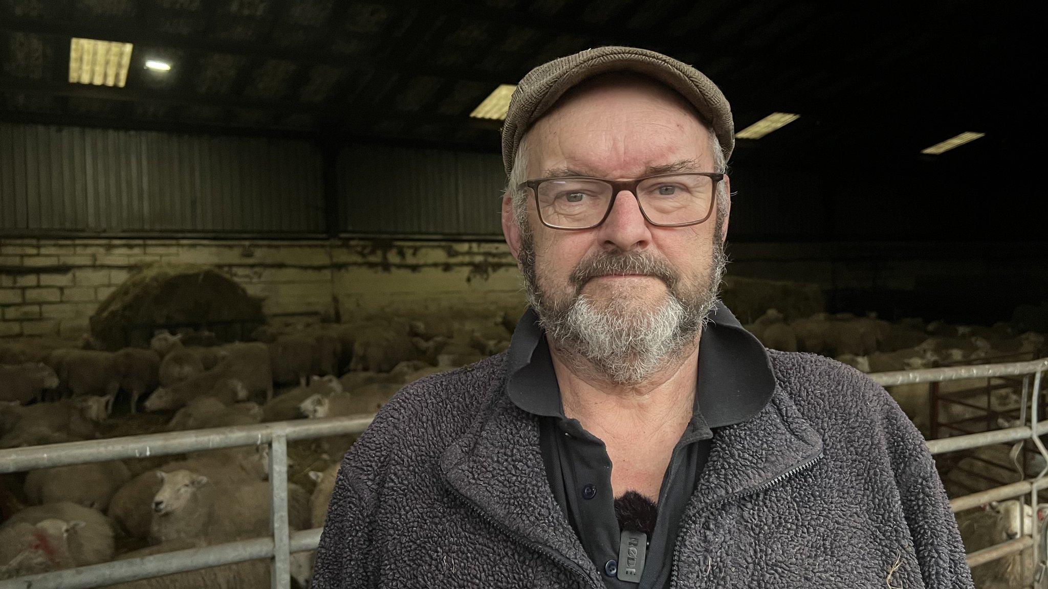 Dave Williams on a farmyard with sheep behind him in a barn