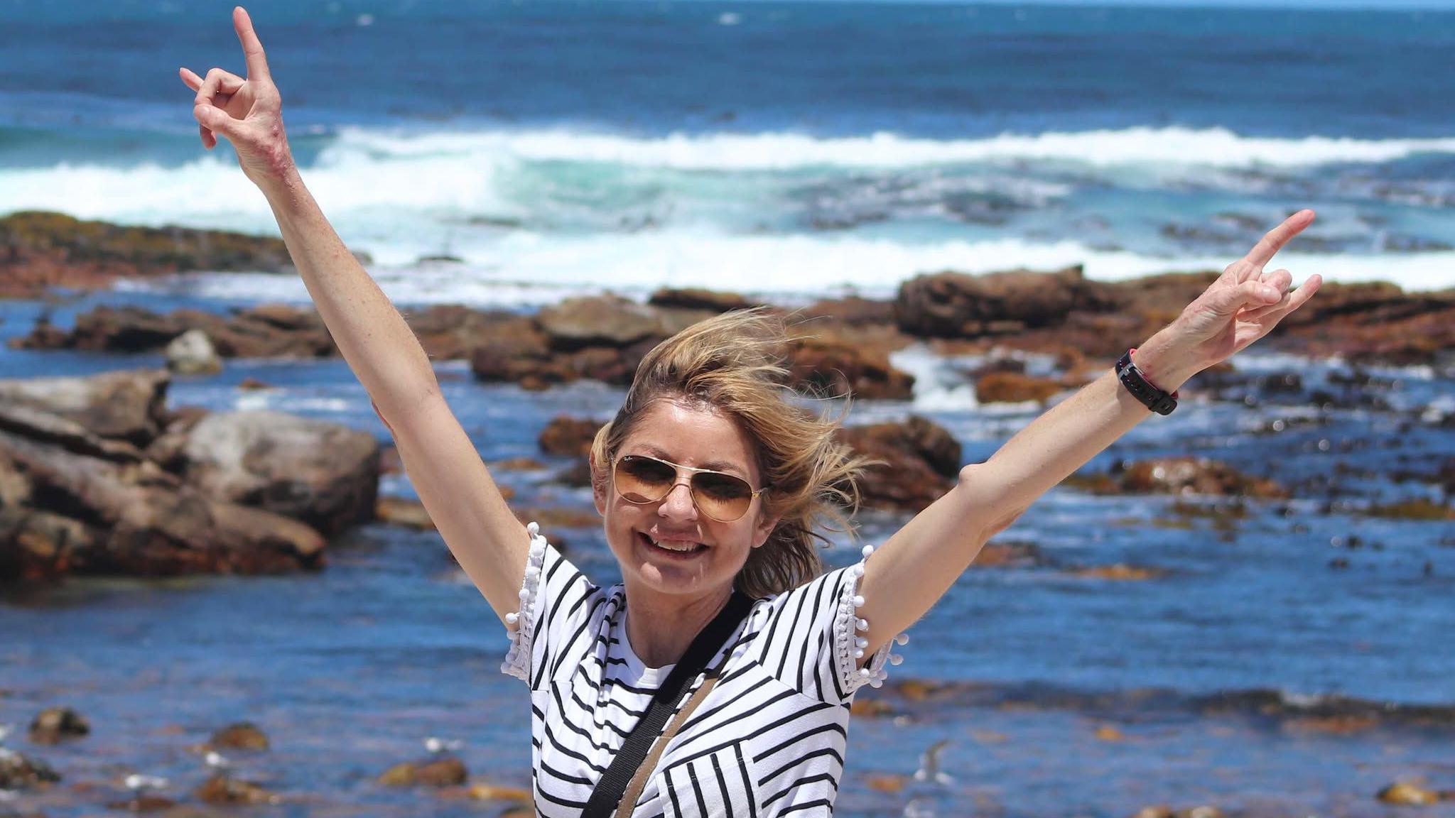 Allison pictured with her arms in the air smiling in the sunshine. She wears glasses and has a navy and white striped top on. 