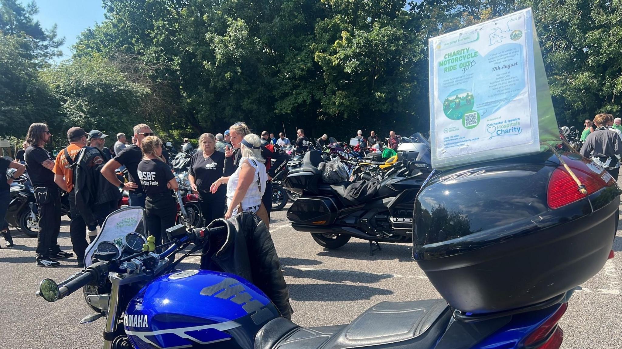 Men and women standing around in a car park, chatting. There are several parked motorbikes. A sign on a bike in the foreground says "Charity motorcycle ride 2025".