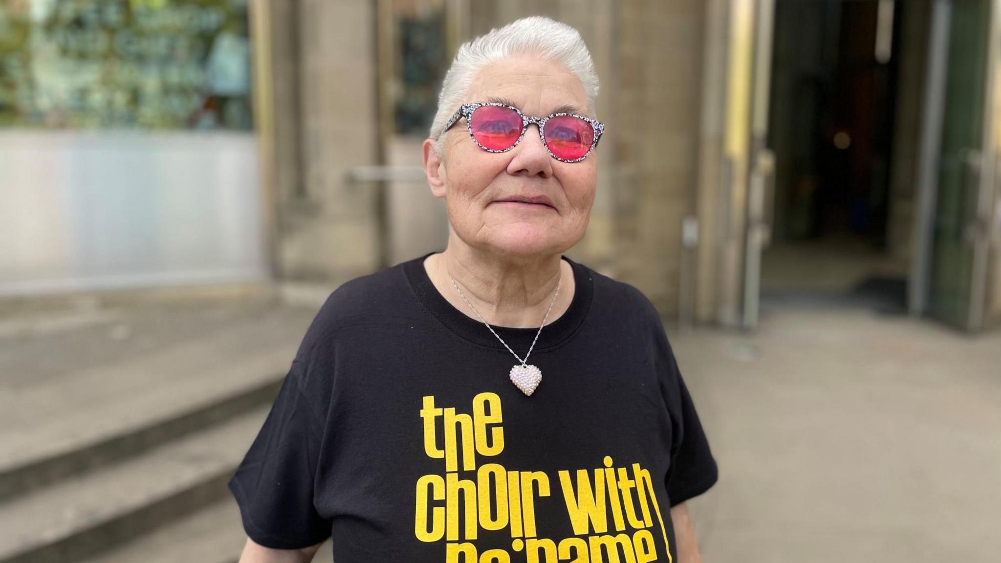 A woman with short white hair and pink glasses smiles to the camera. 