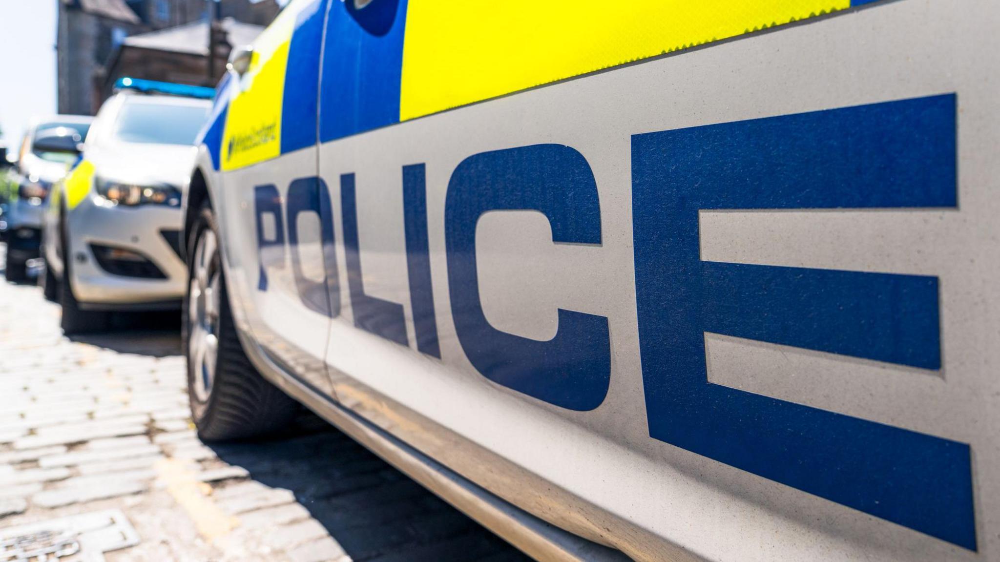 The word 'Police' below the fluorescent decals on a police car on a UK street, with other police vehicles in the distance.