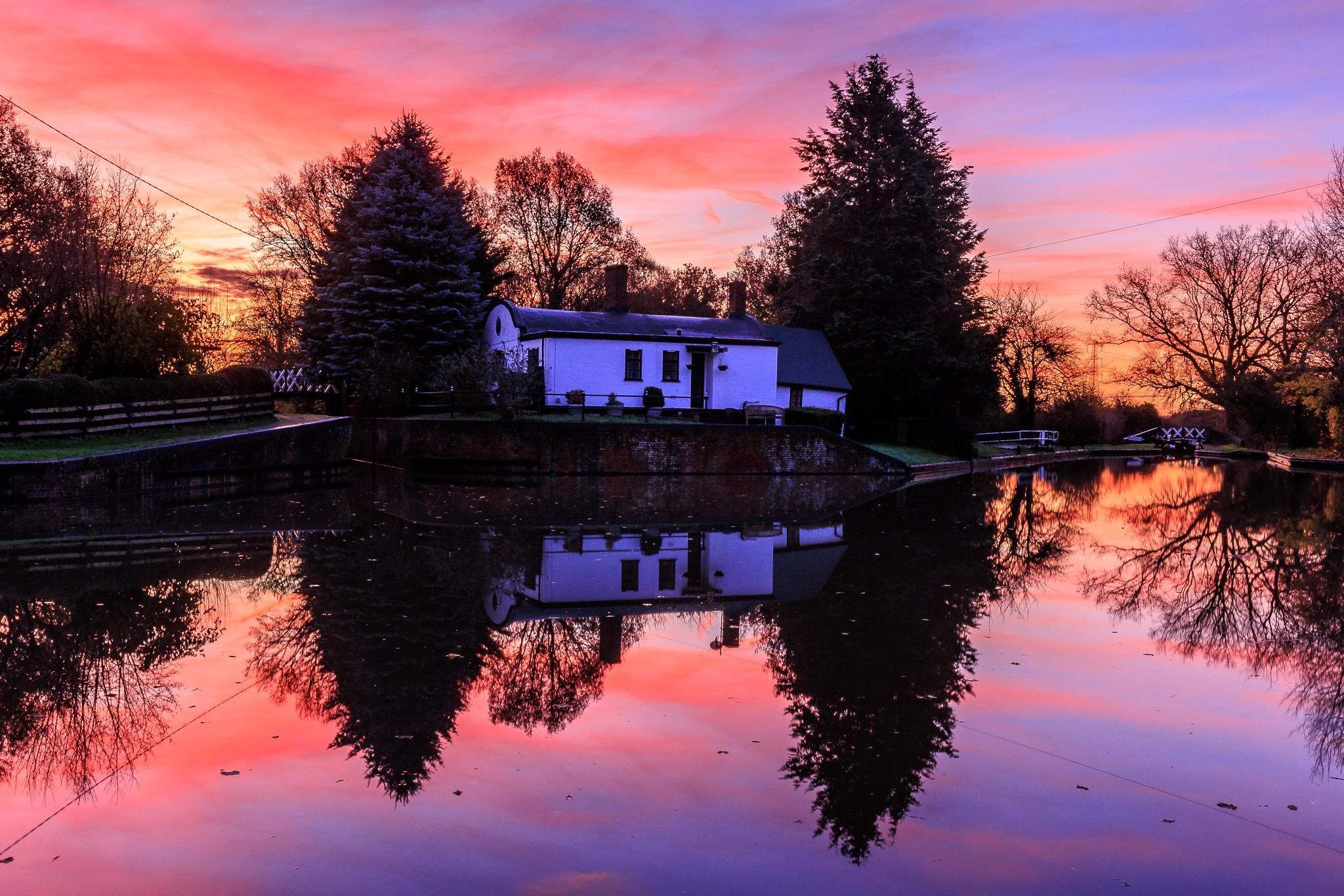 A sunrise of dappled pinks and purples frames a white waterside building next to a canal basin. The scene is perfectly reflected in the still water. 