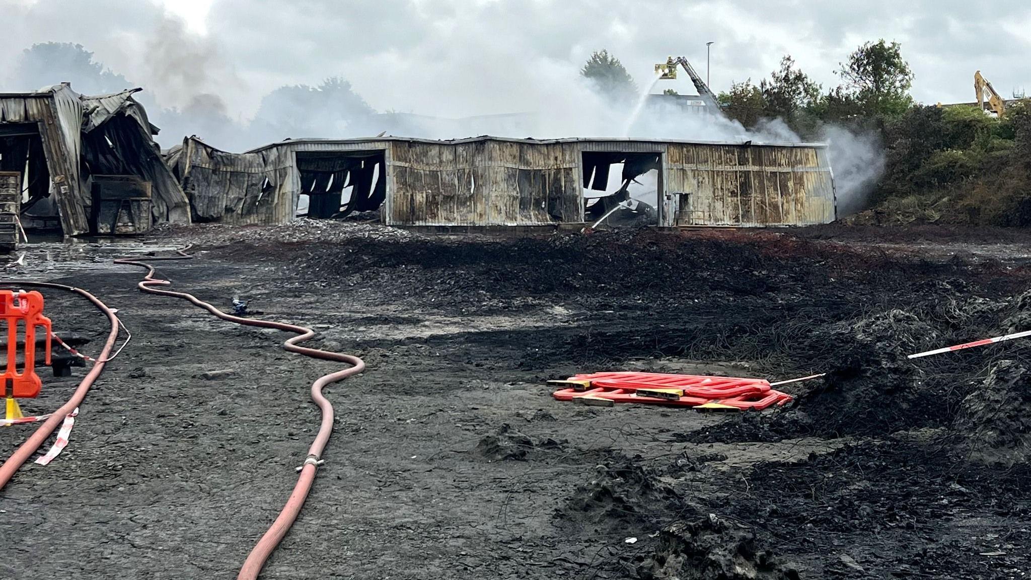 A burnt out building, a large fire hose, red plastic barriers on the ground, smoke in the air, and water being poured on a metal building that is crumpled and collapsed