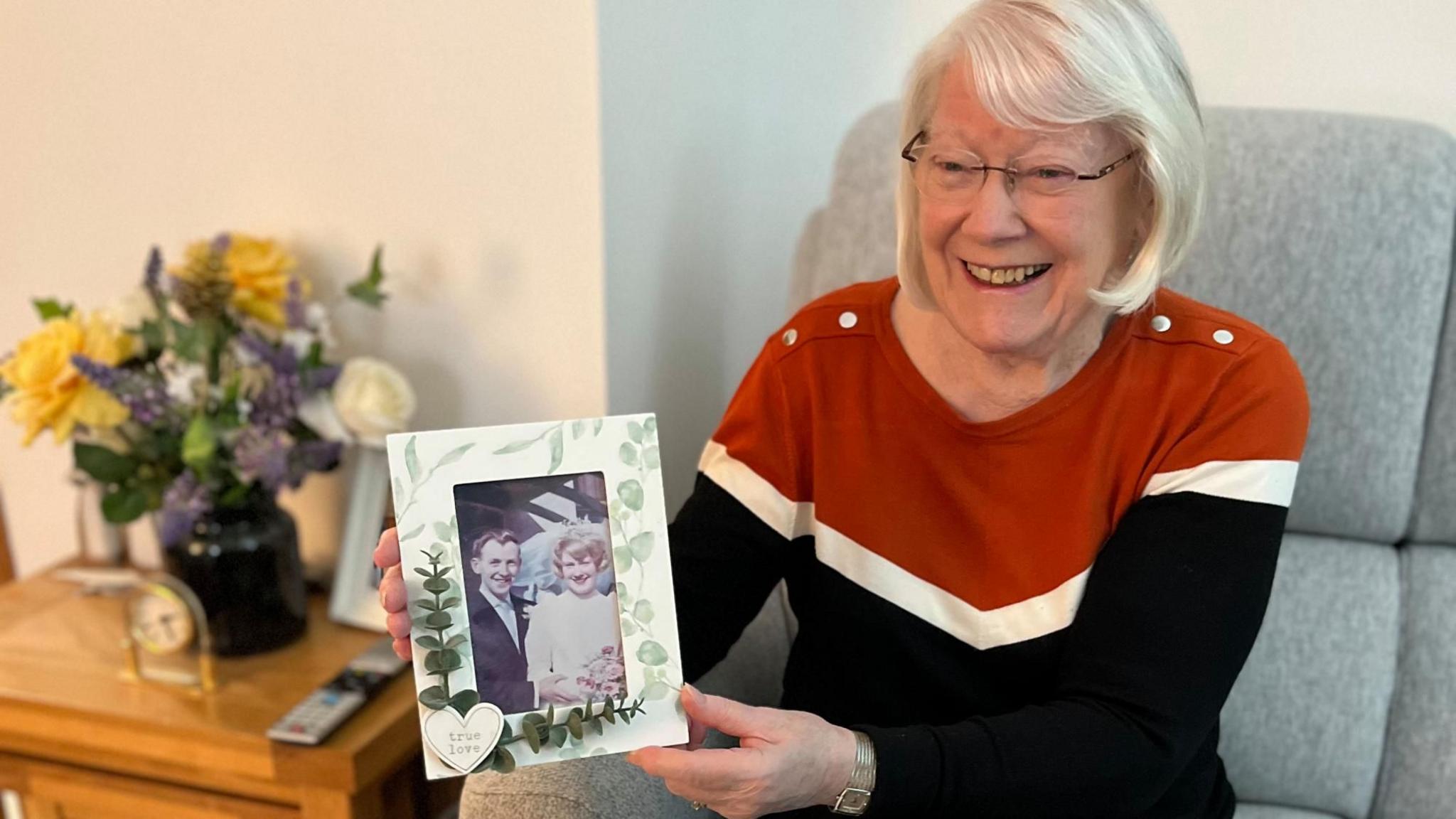 Isobel sits in a chair holding a photo of her wedding day 60 years ago. She has white hair at just less than shoulder length. She is wearing a jumper with red, white and blue stripes. 