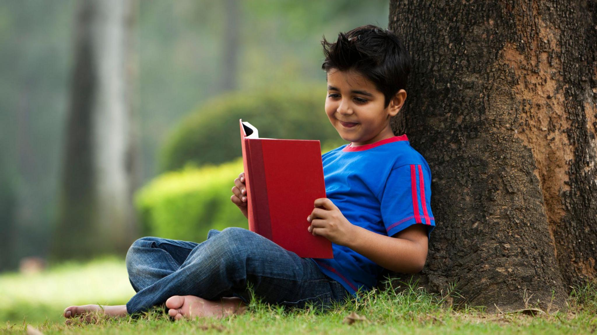 a young boy reading a book under a tree.