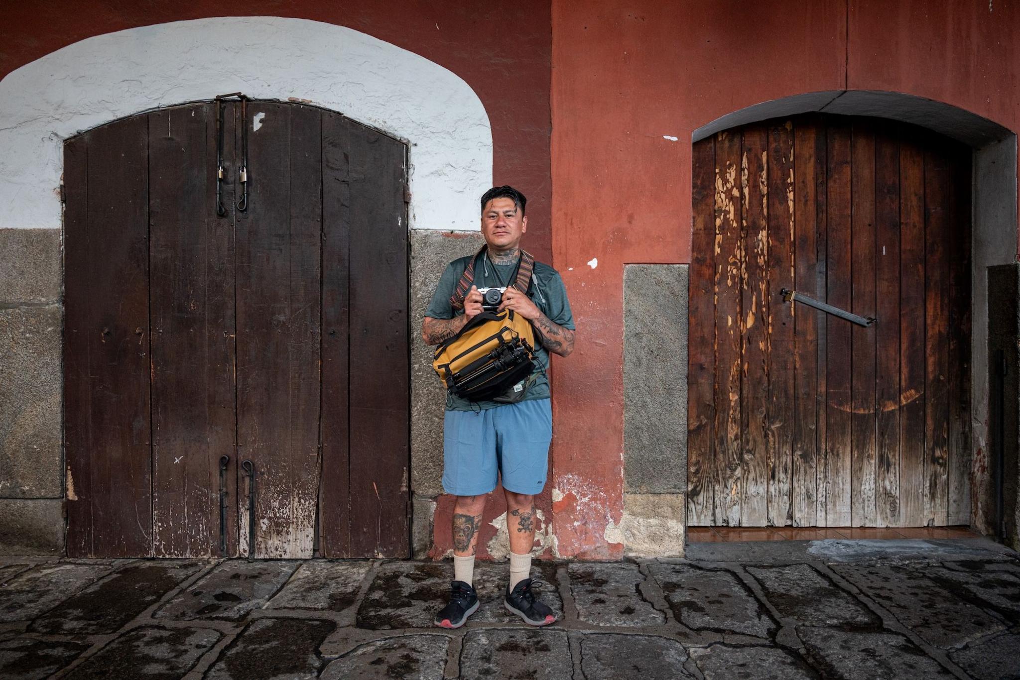 Steven López, wearing shorts and carrying a camera bag, stands in front a building on his way to take photos on his travel across Central America. In his hands, he holds his trusty analogue camera