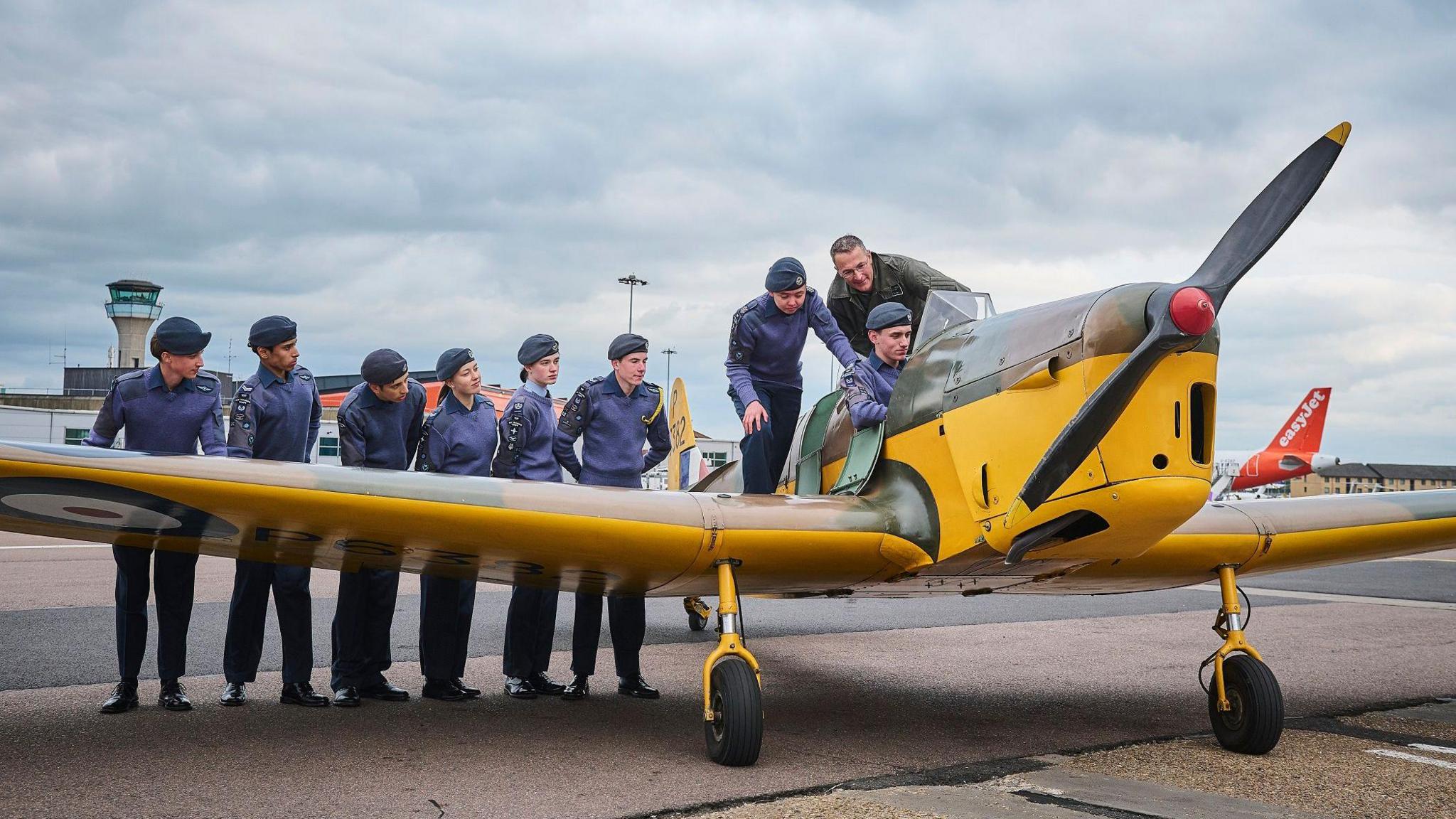 The Miles Magister yellow plane on the forecourt at Luton Airport. A group of eight cadets wearing their purple and black uniform and caps stand behind the left wing, with one sitting in the cockpit and two standing behind him. The pilot wears a green uniform and stands behind the cockpit. The airport control tower is in the background and you can see the orange tail of an easyJet plane.