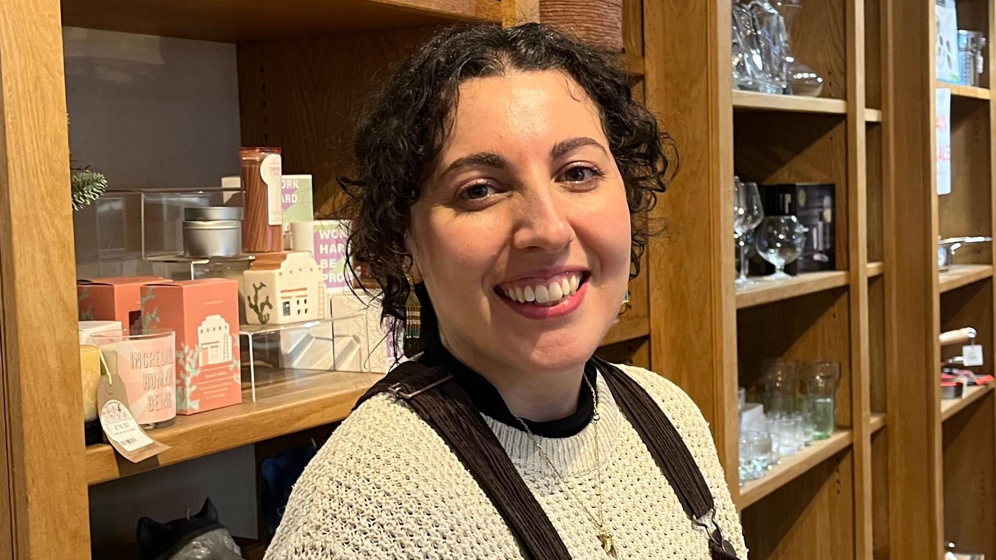 Nicola Hardingham has dark curly hair, cropped to her jawline. She is smiling in the photo, wearing a cream jumper and brown dungarees. She is standing in front of shop shelves filled with items for sale.