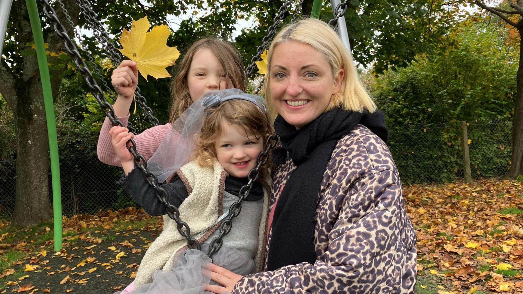 Katy Irwin has blond, shoulder-length hair. She is wearing a leopard-print coat and is smiling at the camera with her two daughters.