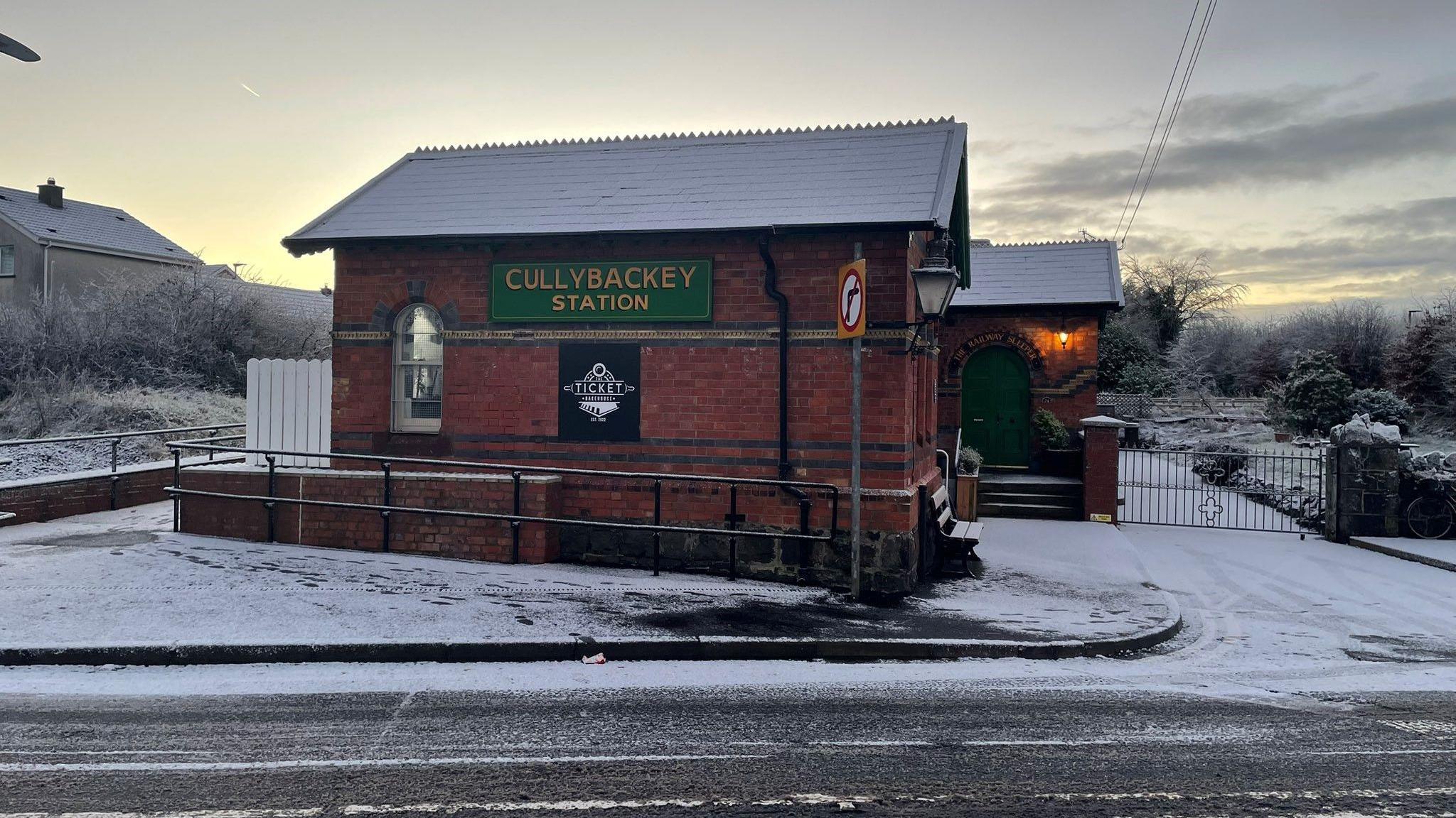 A red brick building, with a green sign on the front reading "Cullybackey Station". The path in front of the building is covered in snow. 