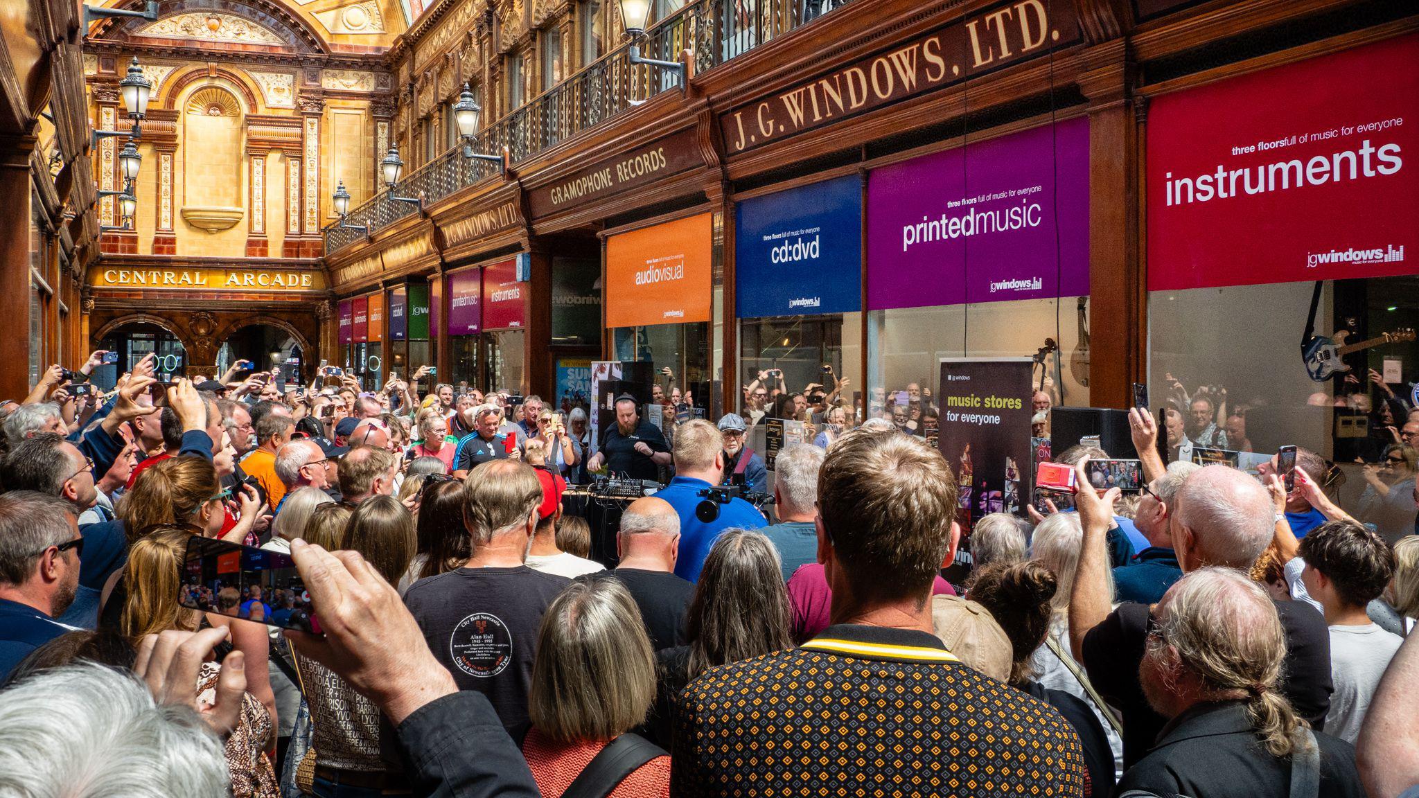 A packed Central Arcade with Lindisfarne playing outside JG Windows. People are taking pictures of the band with their phones.