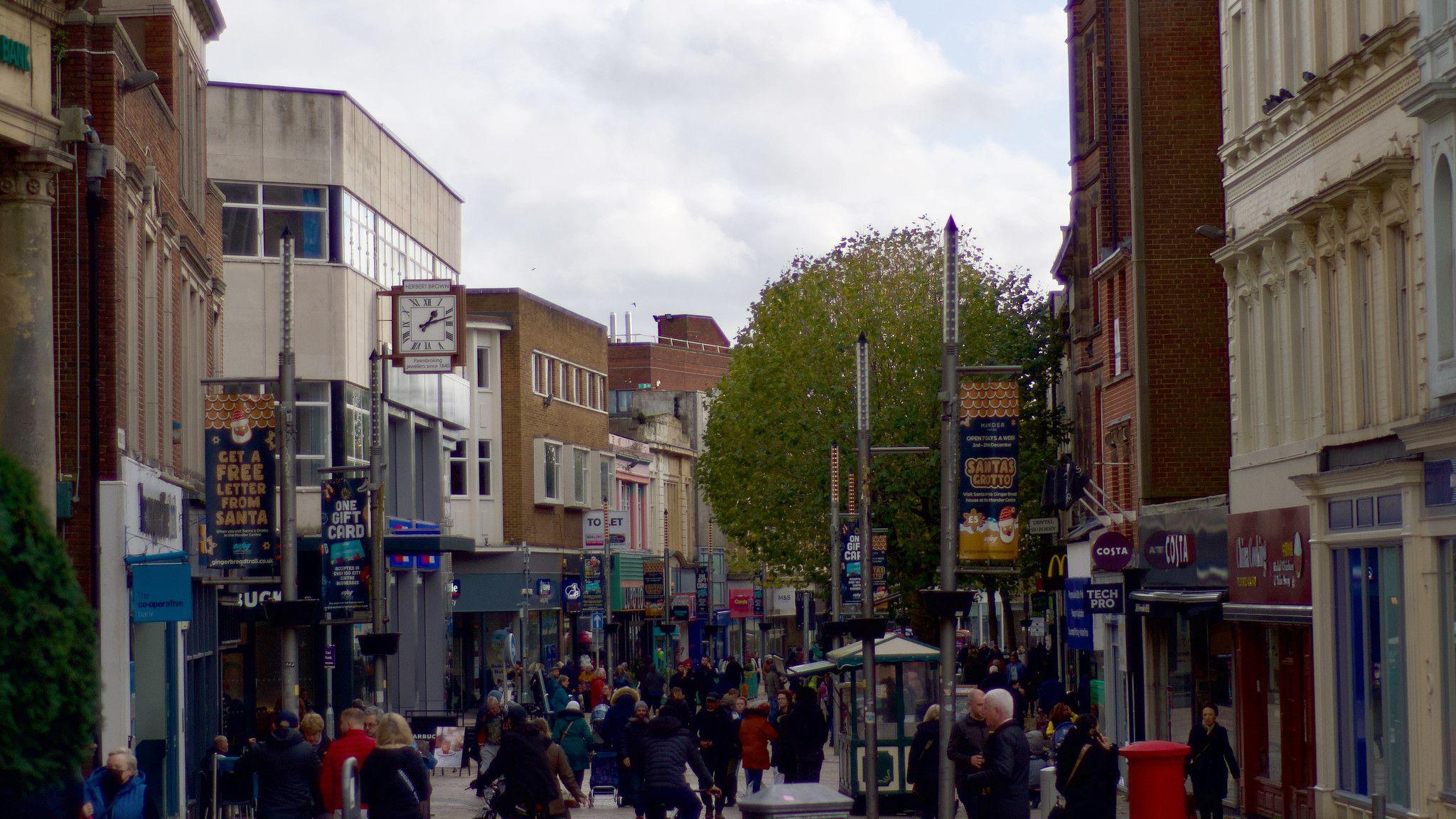 A pedestrianised street full of shoppers. Buildings of varying shades and materials are on both sides.