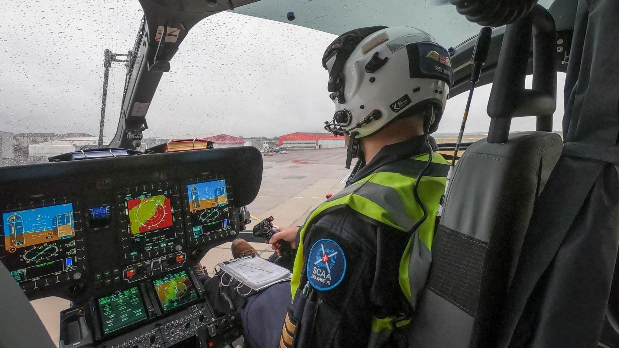 A pilot at the controls in the new helicopter cockpit. He is wearing a high vis jacket, a white helmet and an SCAA badge on his arm. 