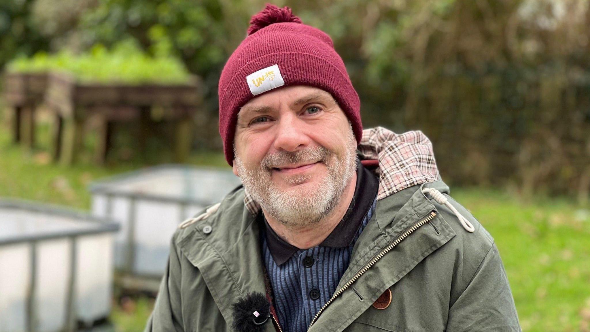 Craig smiles as he looks at the camera. He is wearing a red bobble hat, a blue top and a green coat. He is outdoors in a garden, with some blurred raised planters seen behind him.