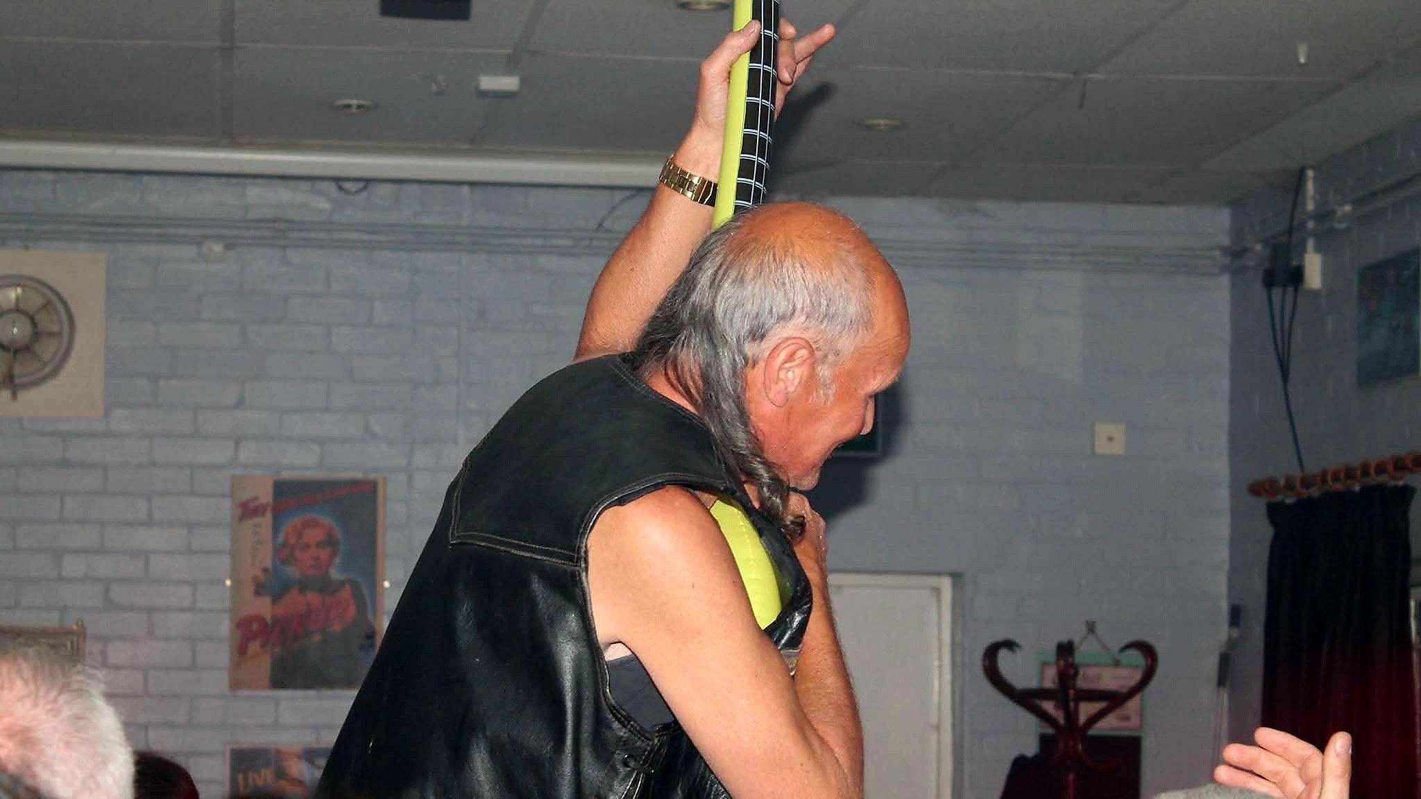 Brian Dean with short white hair, wearing a leather jacket and "playing" a green inflatable guitar, which he is holding vertically. He is in a pub or similar and the hand of an audience member is raised to the right of the picture. There is a hatstand in the background and pictures on a brick wall that has been painted light blue.
