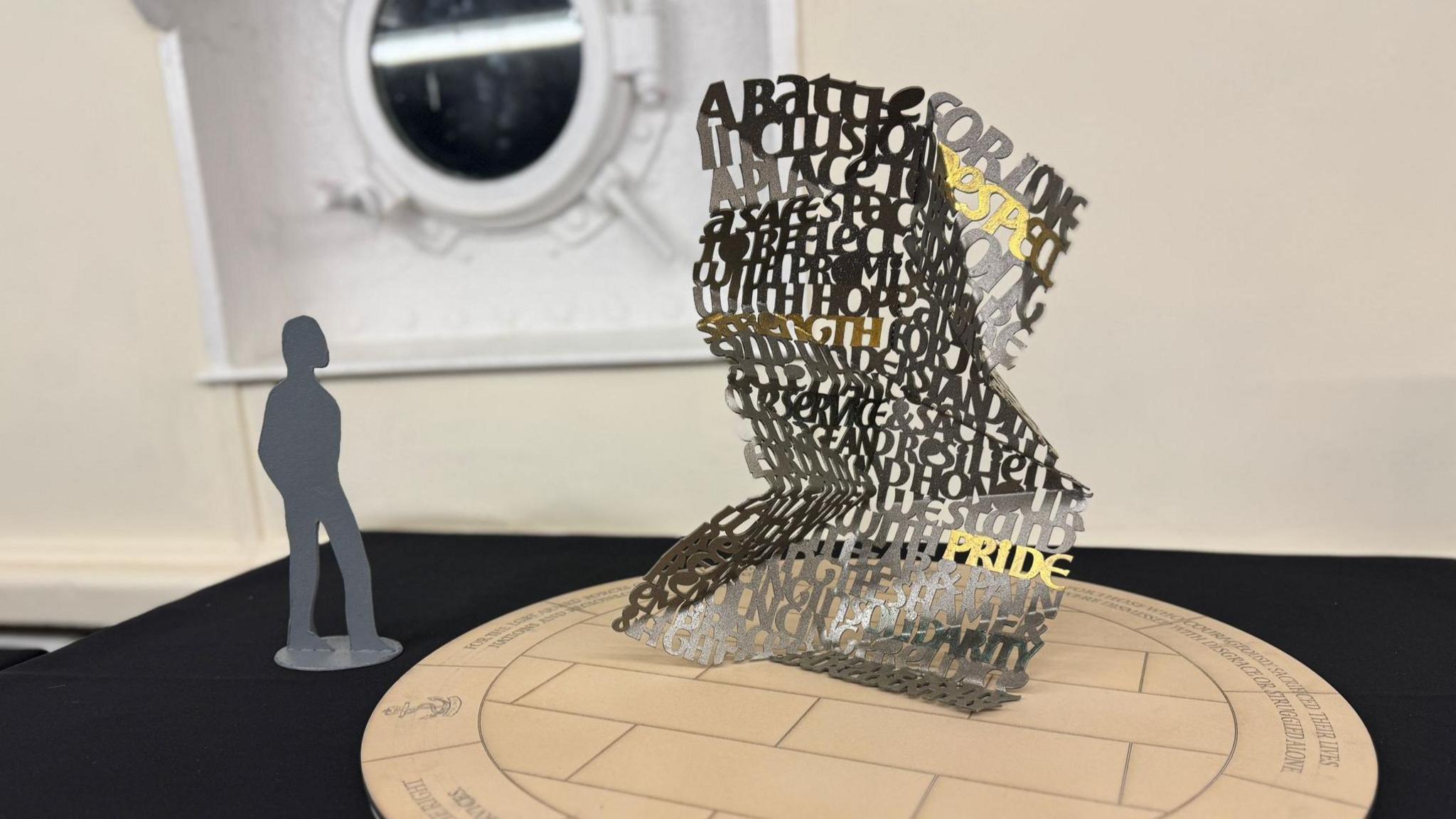 Model of the winning design on a table, next to a small model of a person for scale. The sculpture is approximately twice the height of the person