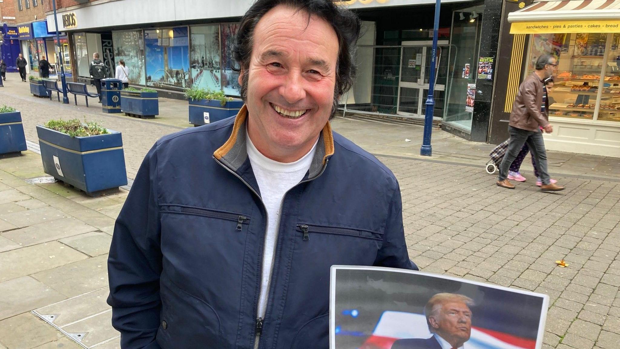 Tony King has black hair in the style of Elvis. He's dressed in a blue jacket and white t-shirt. He's smiling at the camera and holding a picture of Donald Trump.