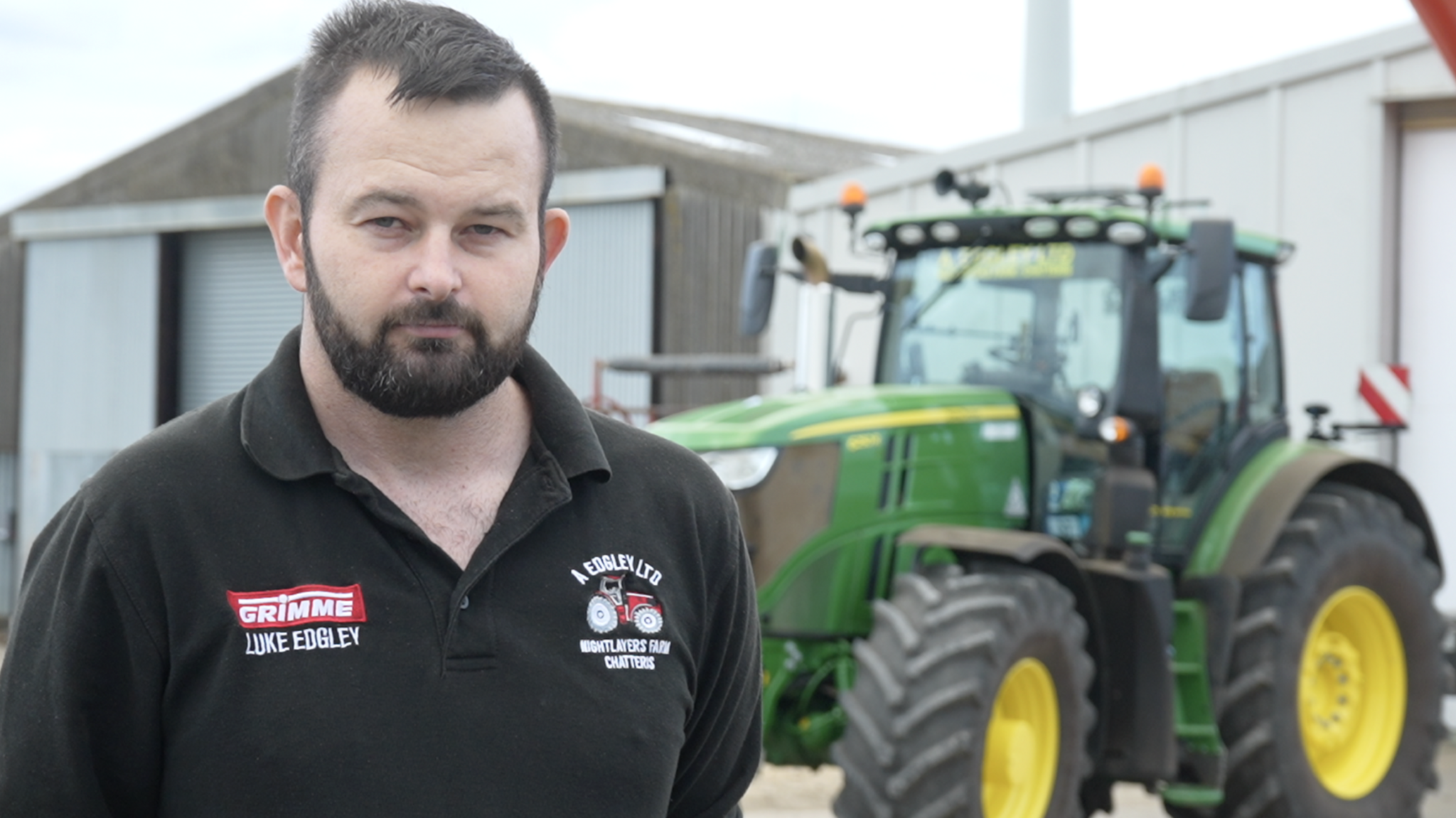 Man wearing black polo shirt stands next to a tractor and looks directly into the camera,