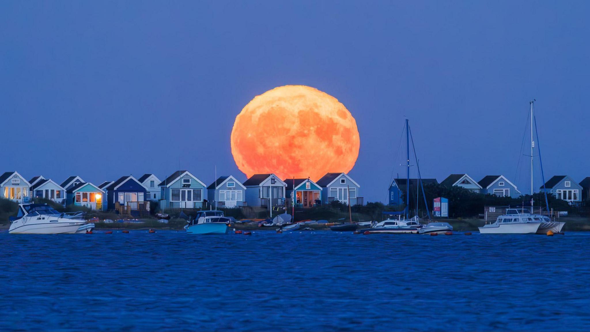 A large orange moon rises in a deep blue sky over colourful beach huts on Mudeford spit. The sky behind is clear some of the colouful beach huts windows are lit up from inside. There are several small boats floating in the foreground on a dark blue sea.