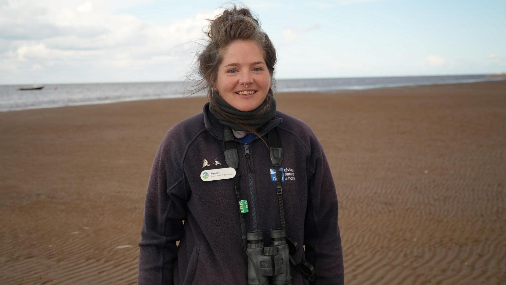 Young woman with hair in a bun and wearing a dark-coloured  jacket and scarf stand on a wide sandy beach with the sea behind. She has a nose ring and the sky is overcast.