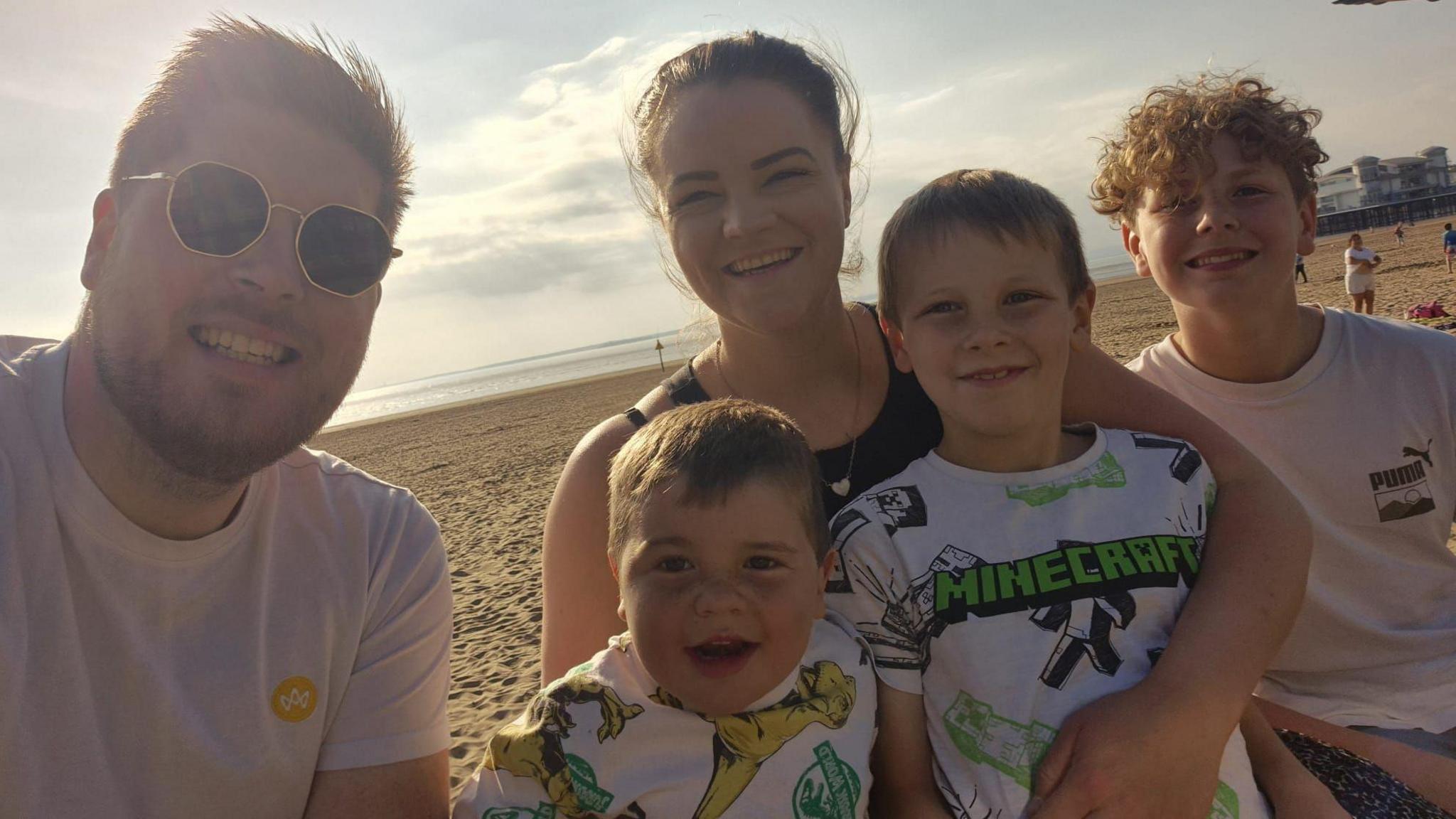 Josh, Shannon and their three sons smile on a beach on a sunny day. There is a wide area of sand behind them and Weston pier is visible in the distance.