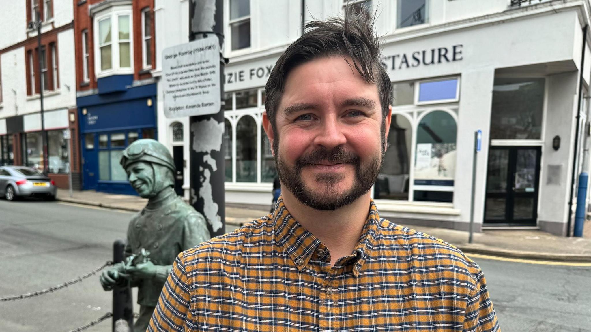 Nick Saunders wears a check yellow, blue and white shirt. He has dark hair and a beard. He stands near the green statue for Formby by a roadside. He smiles.