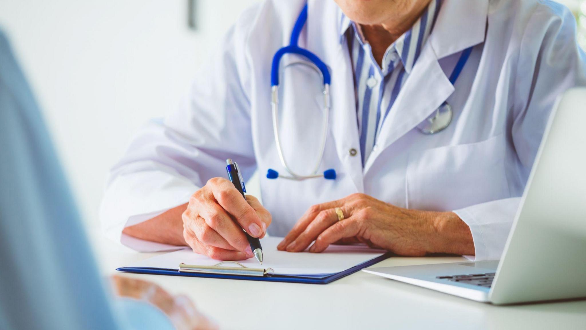 A person wearing a white coat with a blue stethoscope around their neck holds a pen while writing on a clipboard. They are shown from the shoulders down and is sat at a white table with a laptop open in front of them. A second person with their back to the camera is on the left hand side of the image and slightly blurred.