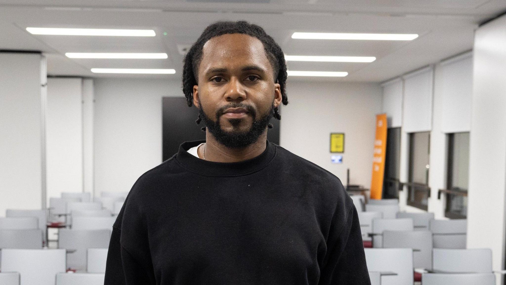 Levander Walters is seen in a black jumper standimg in an empty conference room, facing the camera.