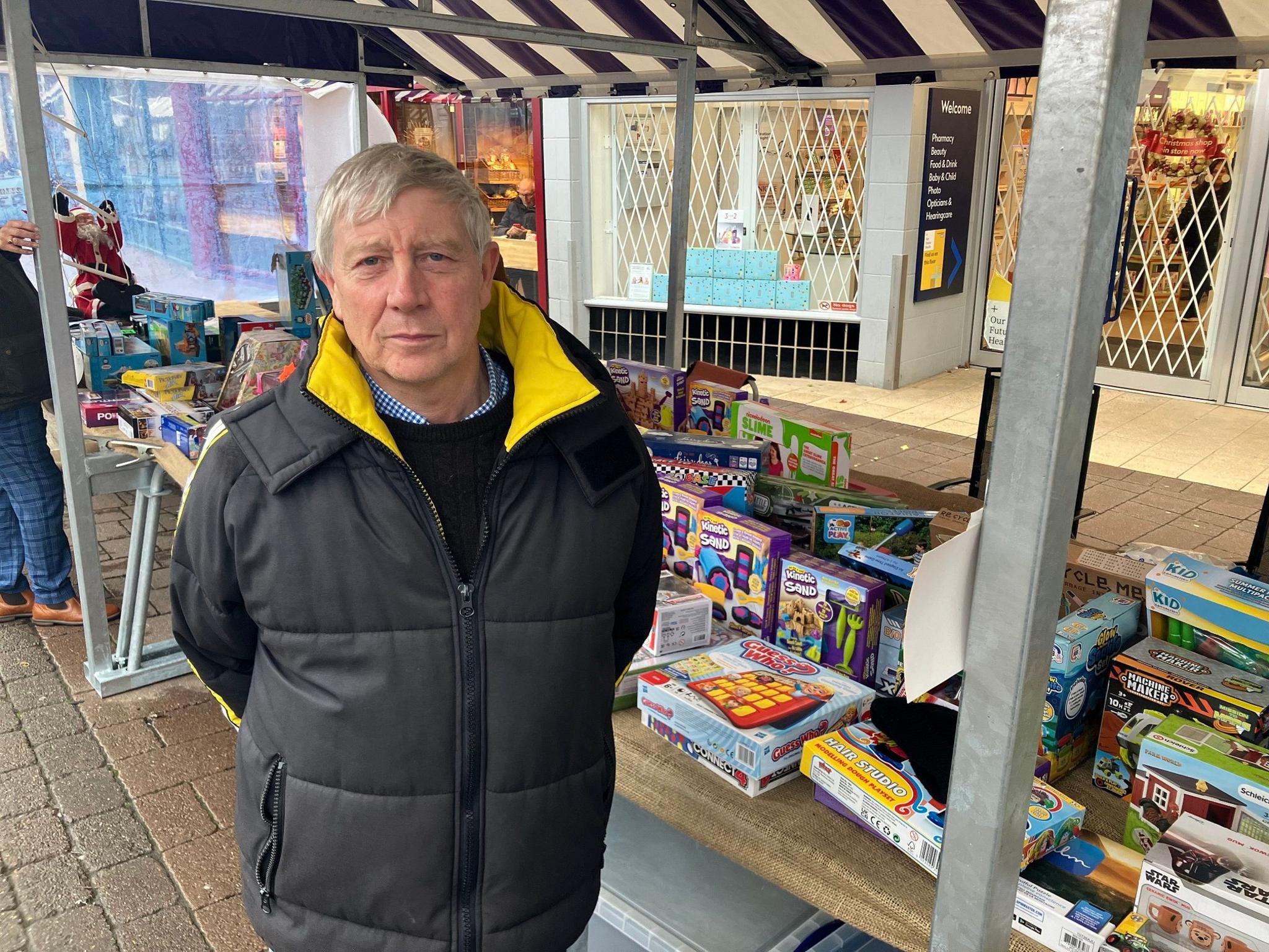 A man in a black coat with a smart yellow trim stood by a market stall selling toys and games