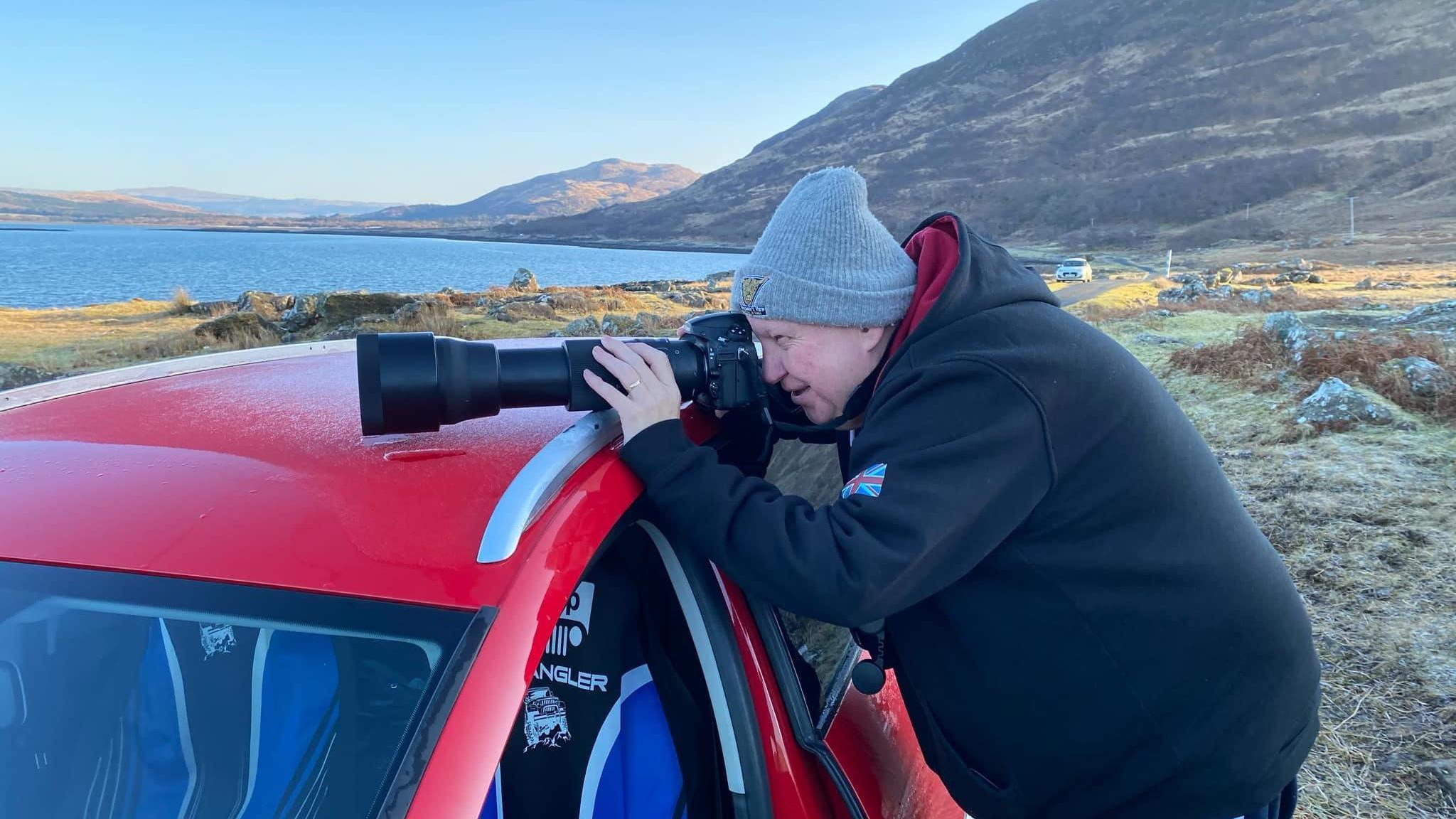 Colin Morrison, wearing a jacket and grey woolly hat, leaning on a red car, with a camera with a long lens propped on the roof. He is leaning into the camera to take a picture. There are hills and a loch behind him.