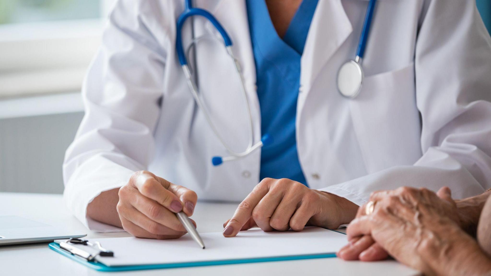 A doctor, wearing scrubs and a white coat, sitting at a desk with a clipboard on it. They are holding a pen. The hands of an older person are also visible in the picture.