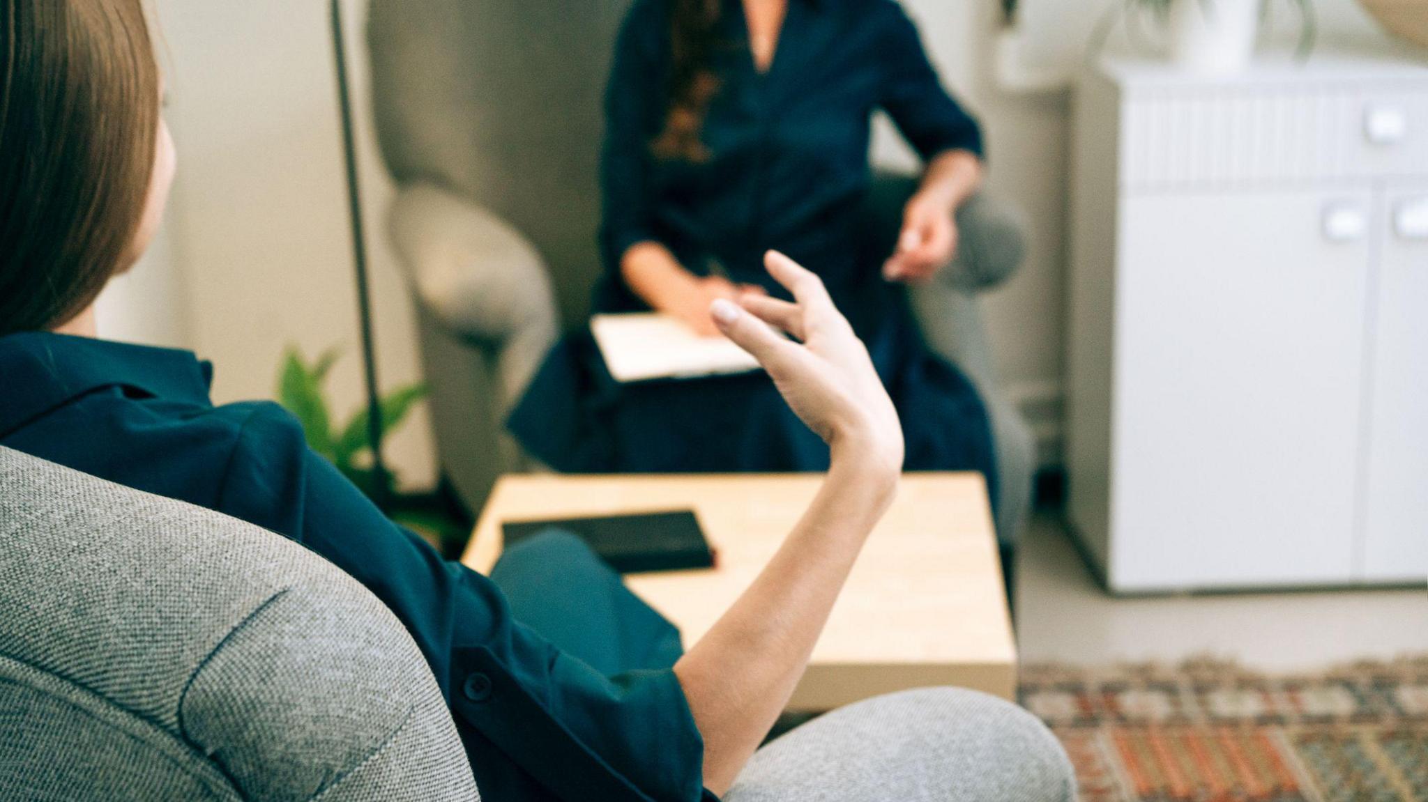 Two women are sitting on chairs talking to each other. They are both wearing blue shirts and have dark hair.