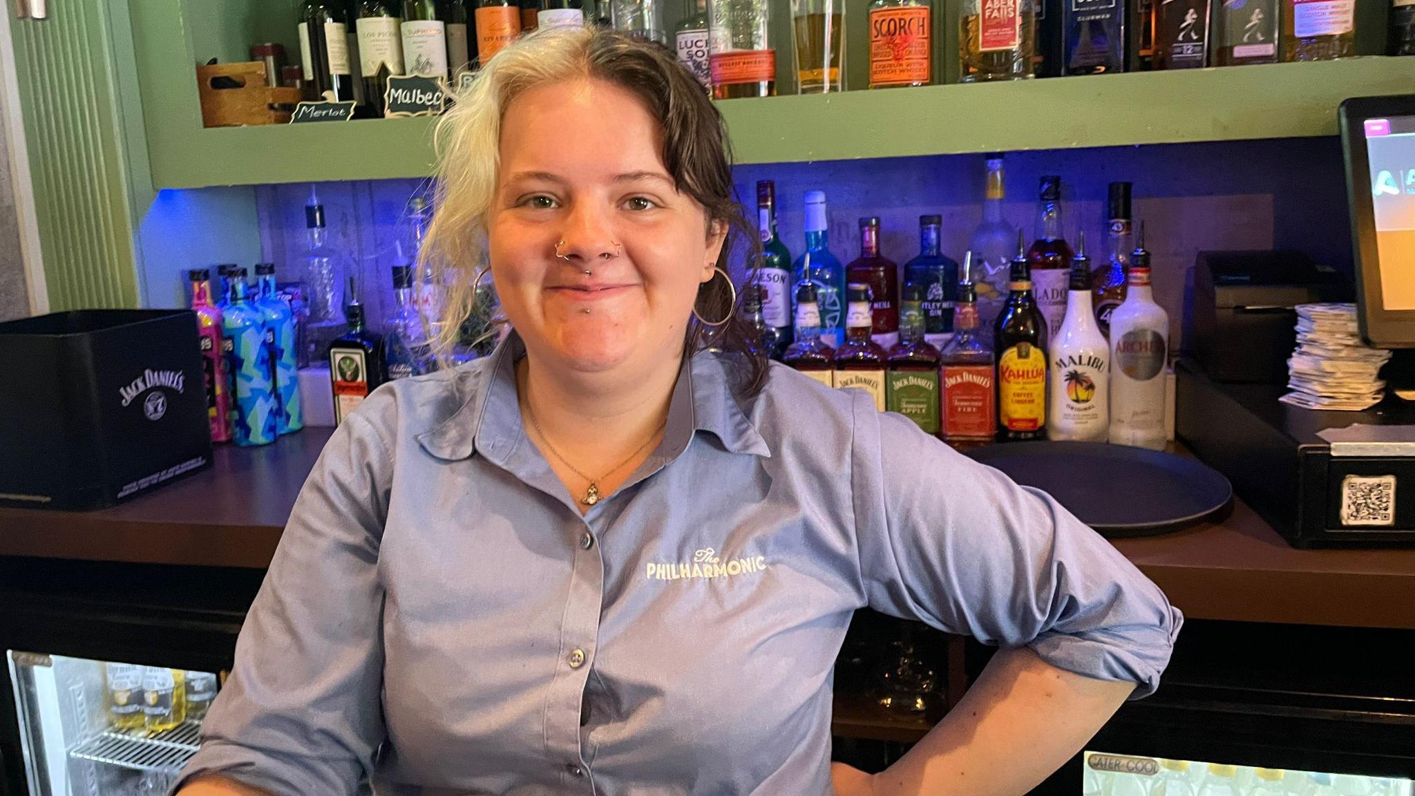 Meg wearing a blue branded shirt at the Philharmonic bar. She has blonde and black hair split down the middle and is wearing hoop earrings with several piercings around her nose. There are bottles of alcohol behind her at the bar with a fridge on her right.