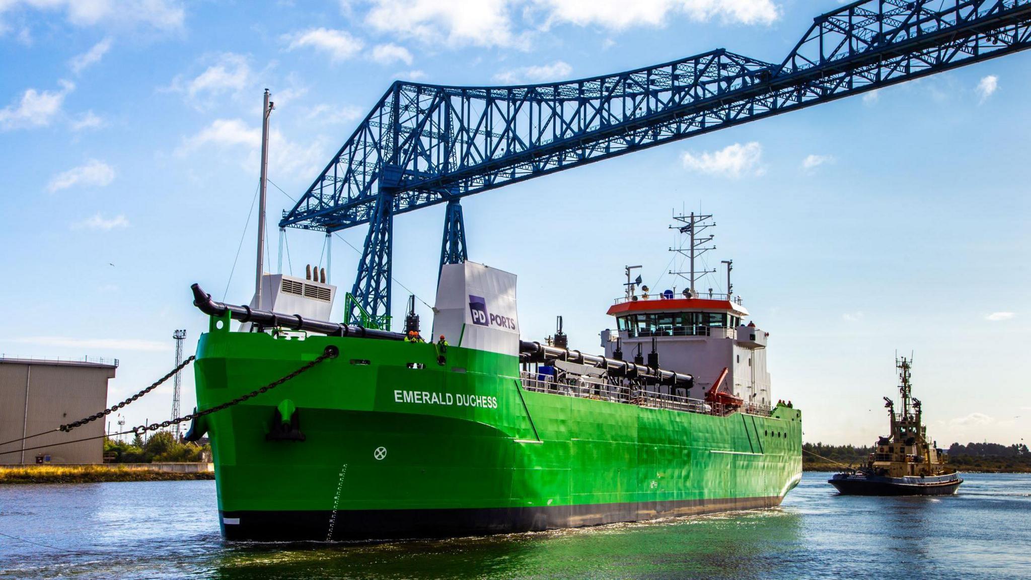 Three quarter side view of a dredger named Emerald Duchess. The green vessel, with a tug behind it, is passing under the Middlesbrough Transporter Bridge.