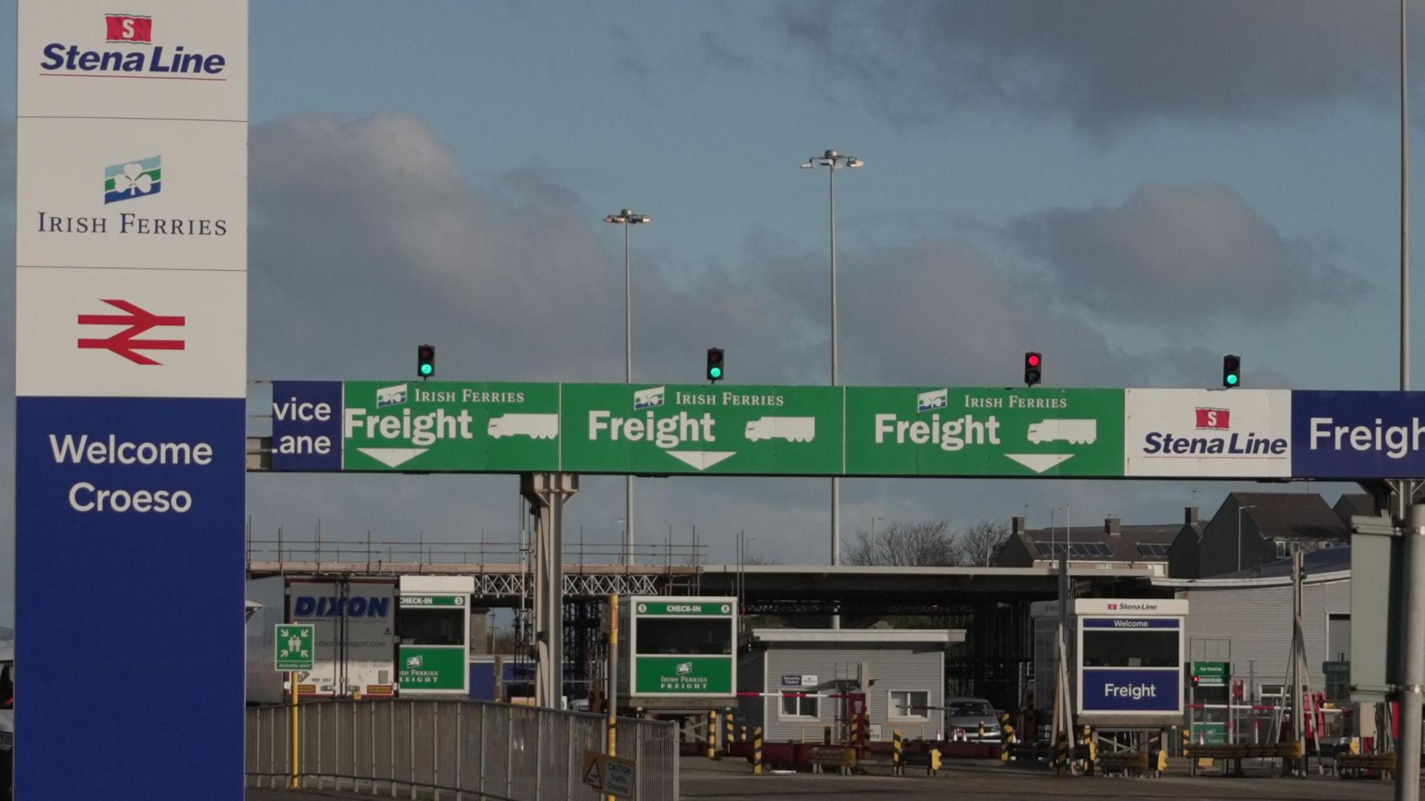 Three green signs above different lanes directing freight traffic on to Irish Ferries service to Dublin are seen at the Port of Holyhead in Holyhead. To the left of these is a large blue and white 'Welcome' sign with the various transport companies listed. There are no vehicles driving towards the barriers.