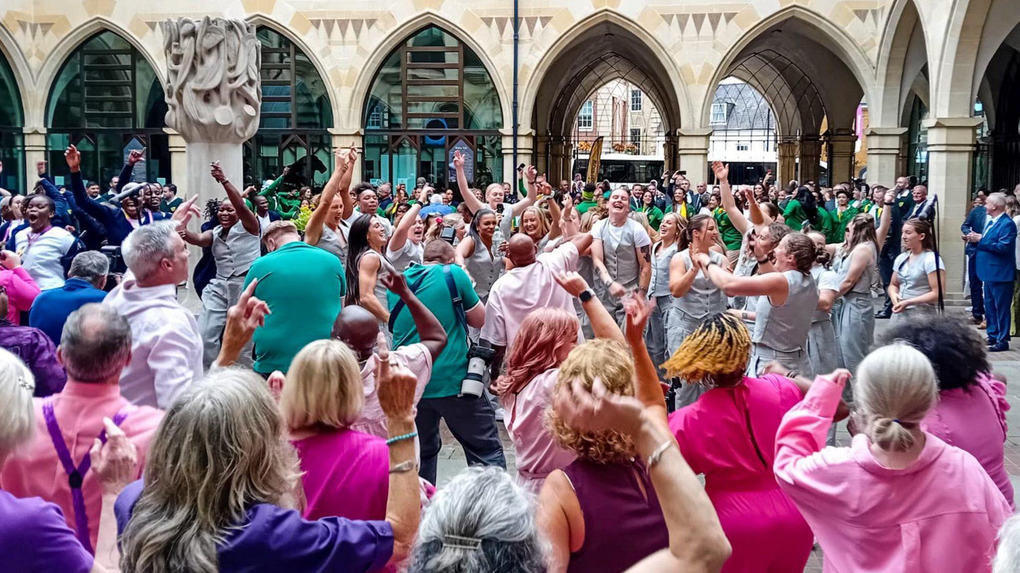 A colourful scene of a large group of people singing and dancing in the courtyard of Northampton's Guildhall.