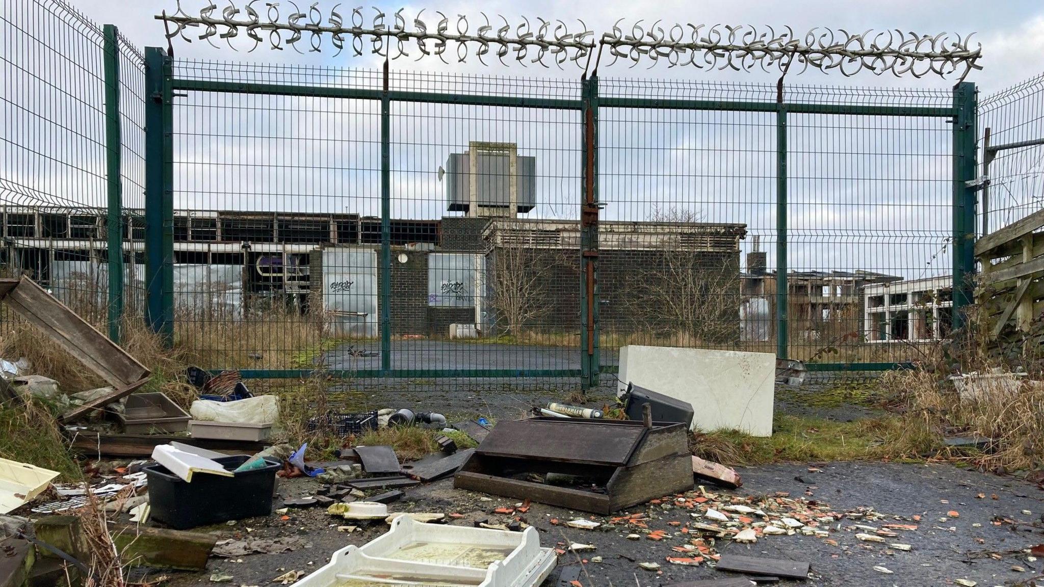 A discoloured fridge door lying on its back and a broken brown, wooden bit of school furniture are among the detritus dumped outside the green metal gates of the former school. 