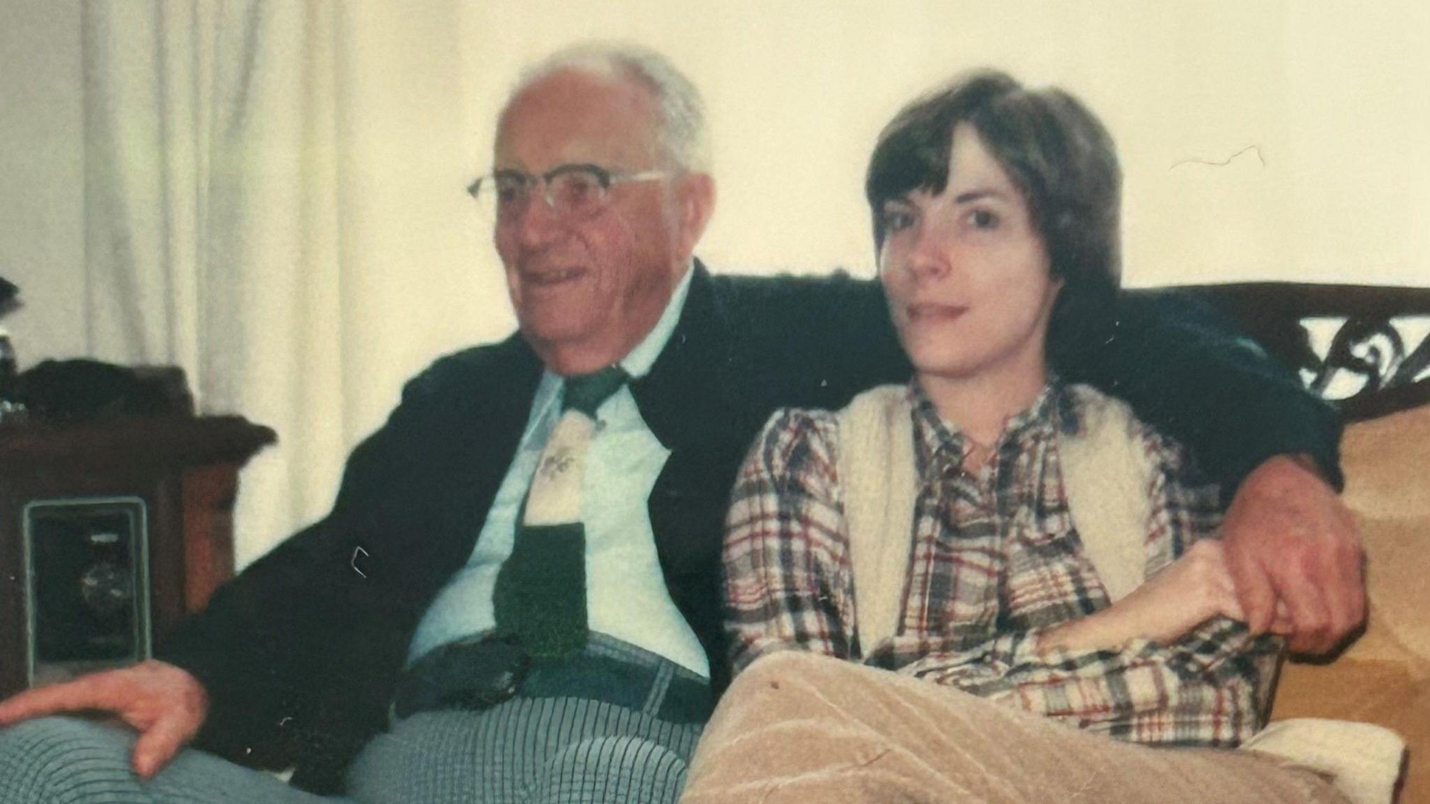 George Clarke is sitting with his daughter Trudy Johnson in a photograph dating from the 1980s. He is probably in his 70s and is wearing a suit and has his arm around Trudy who is in her 20s and wearing a patterned shirt and white waistcoat.