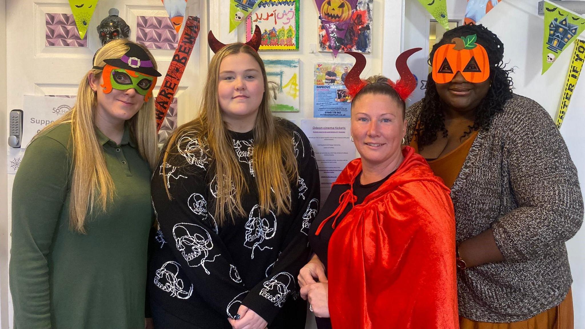 Four women standing in front of a display board with children's drawings on it and they are all wearing Halloween fancy dress. The lady on the left, with long blonde hair and a green top, wears a witches-style pair of glasses. The woman next to her, also with long blonde hair, has a black top with white skulls on it. Next to her is a woman with dark hair pulled back, a pair of novelty red horns on her head and a black cape. On the right is a woman with curly black hair to her shoulders, pumpkin-style glasses on her face and wears a grey cardigan over a orange/brown dress.